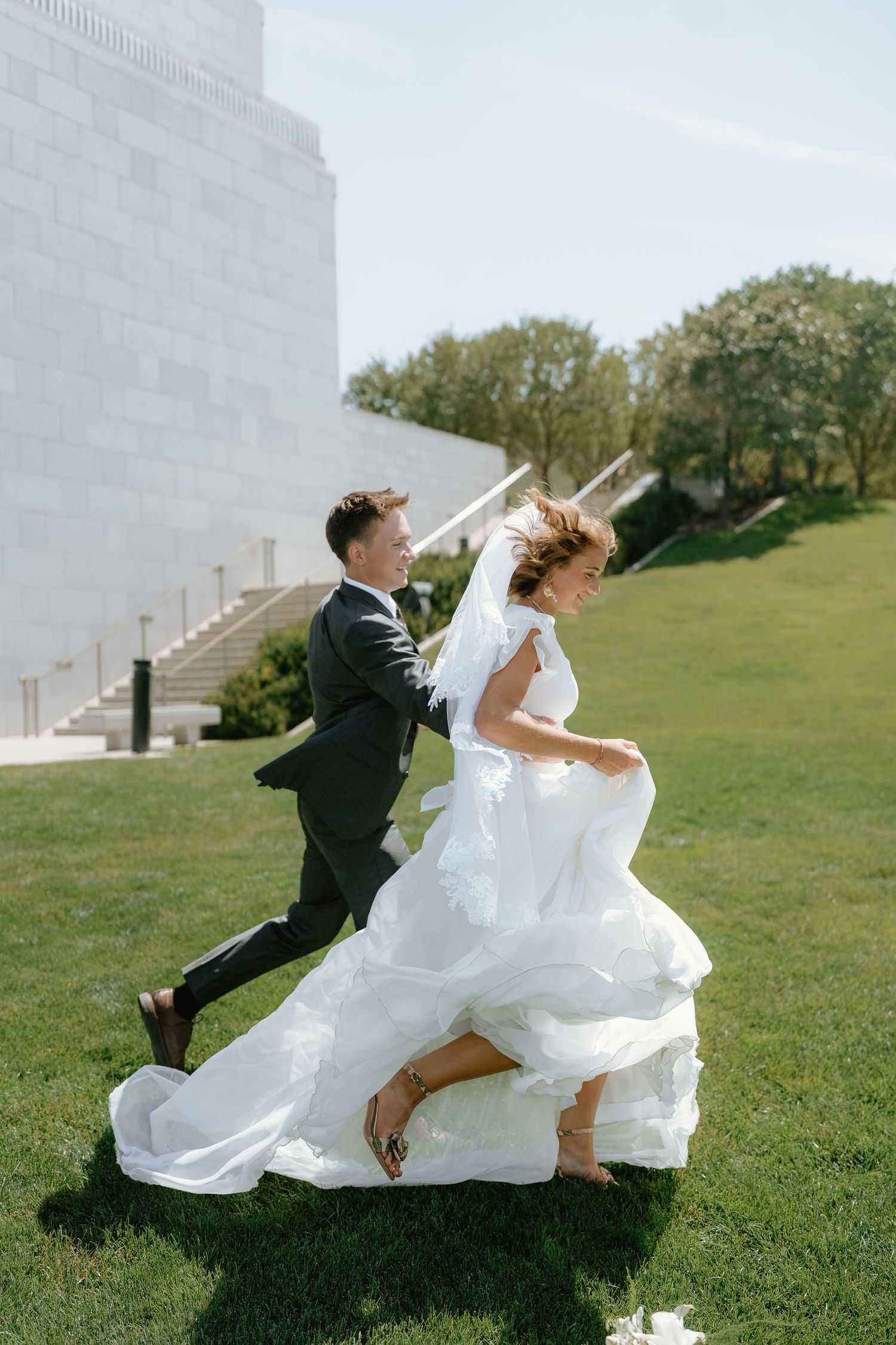 Bride and groom running and laughing on green lawn outside white wedding venue on sunny day.