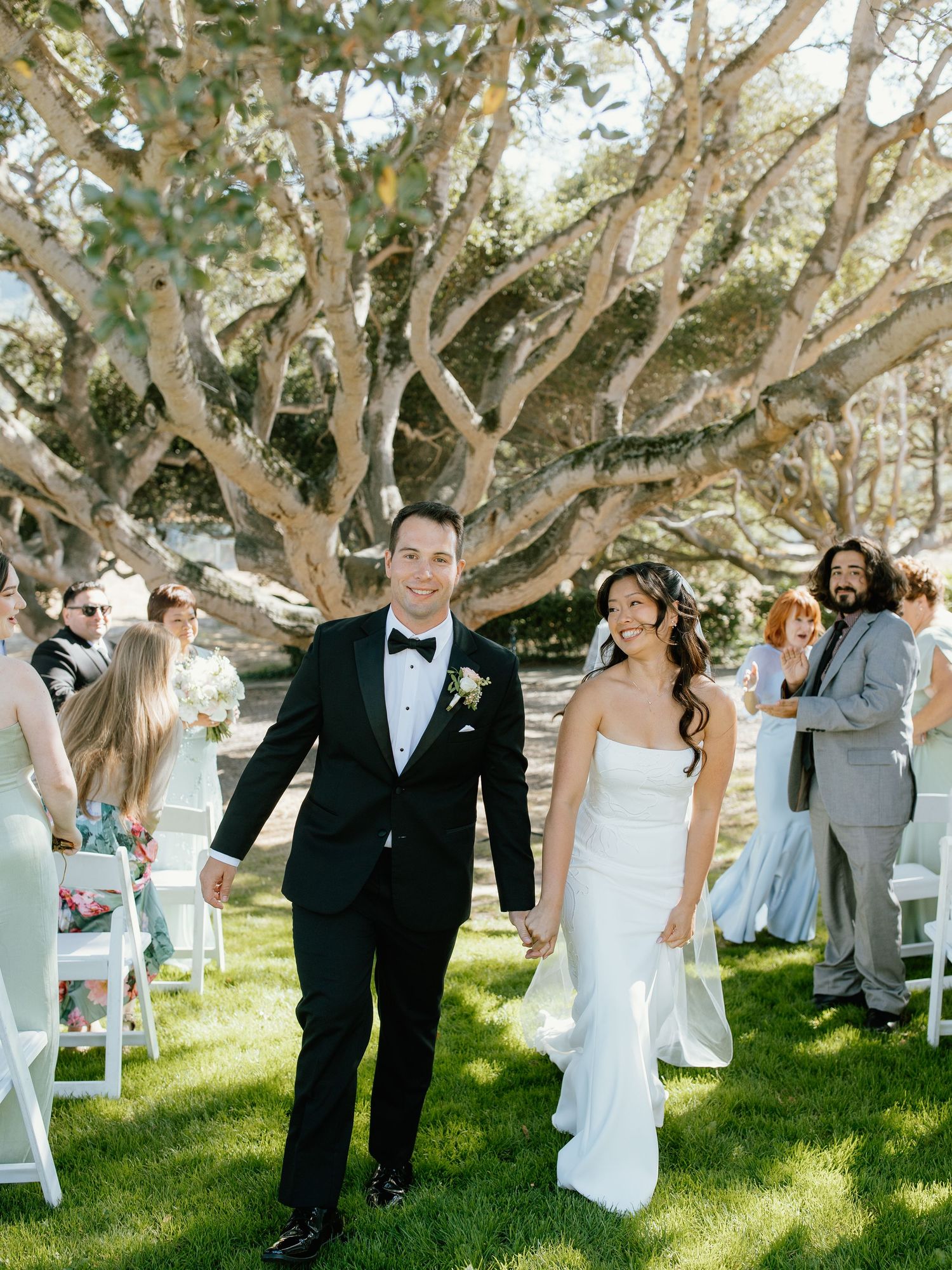 Bride and groom walk down outdoor aisle holding hands under sprawling tree with wedding guests seated nearby.