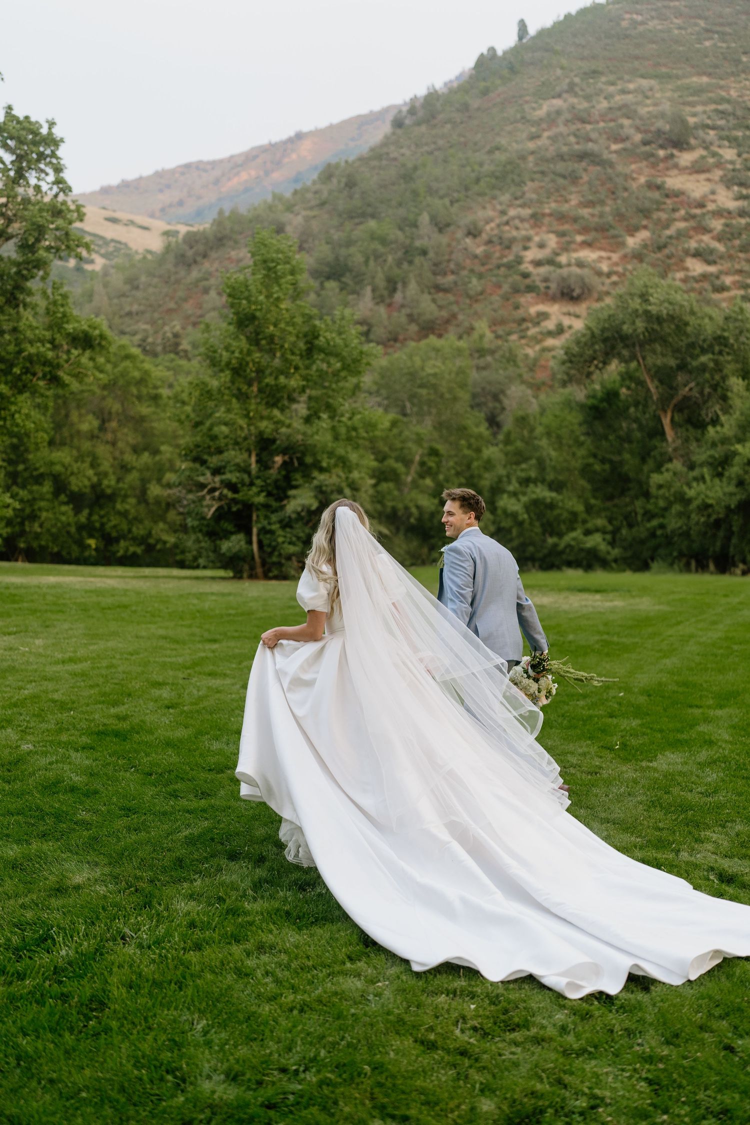 Bride in flowing gown and long veil walks with groom across lush green mountain meadow.