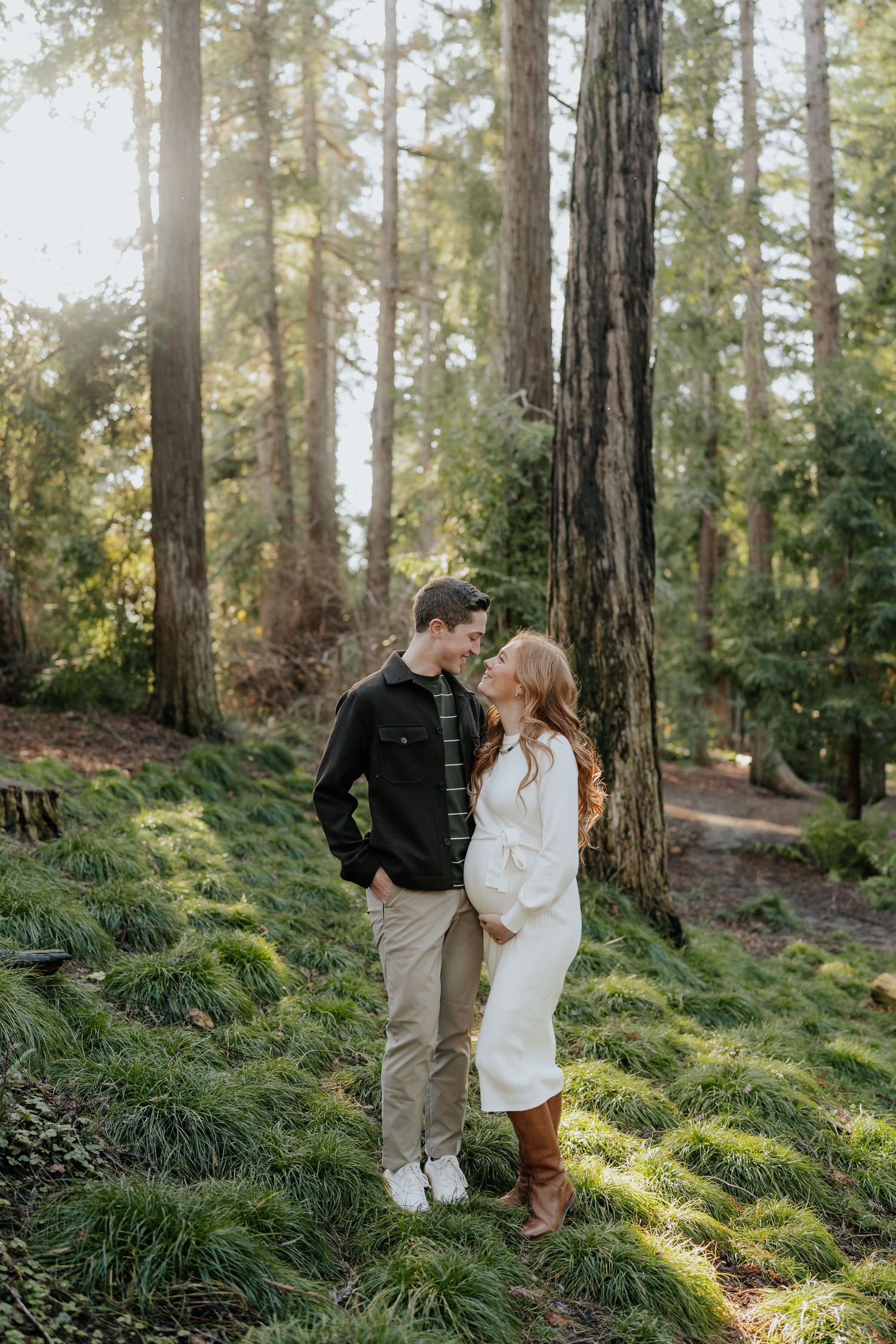 Couple gazes lovingly at each other in a sunlit redwood forest with lush green ground cover in Davis, California.