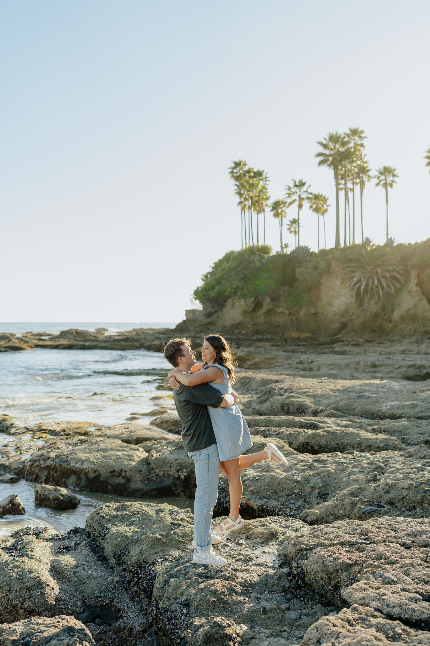 Couple embracing and laughing on rocky beach with palm trees and ocean backdrop in Laguna Beach, California.