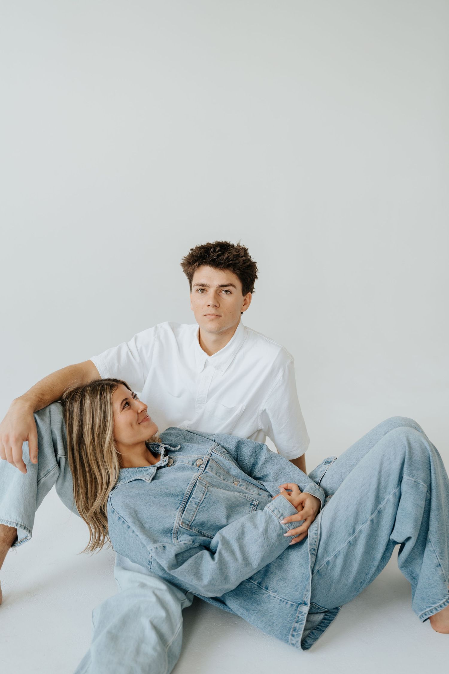 Couple in matching denim outfits relaxing on white studio floor in casual poses at Studio Elevn in Salt Lake City, Utah.