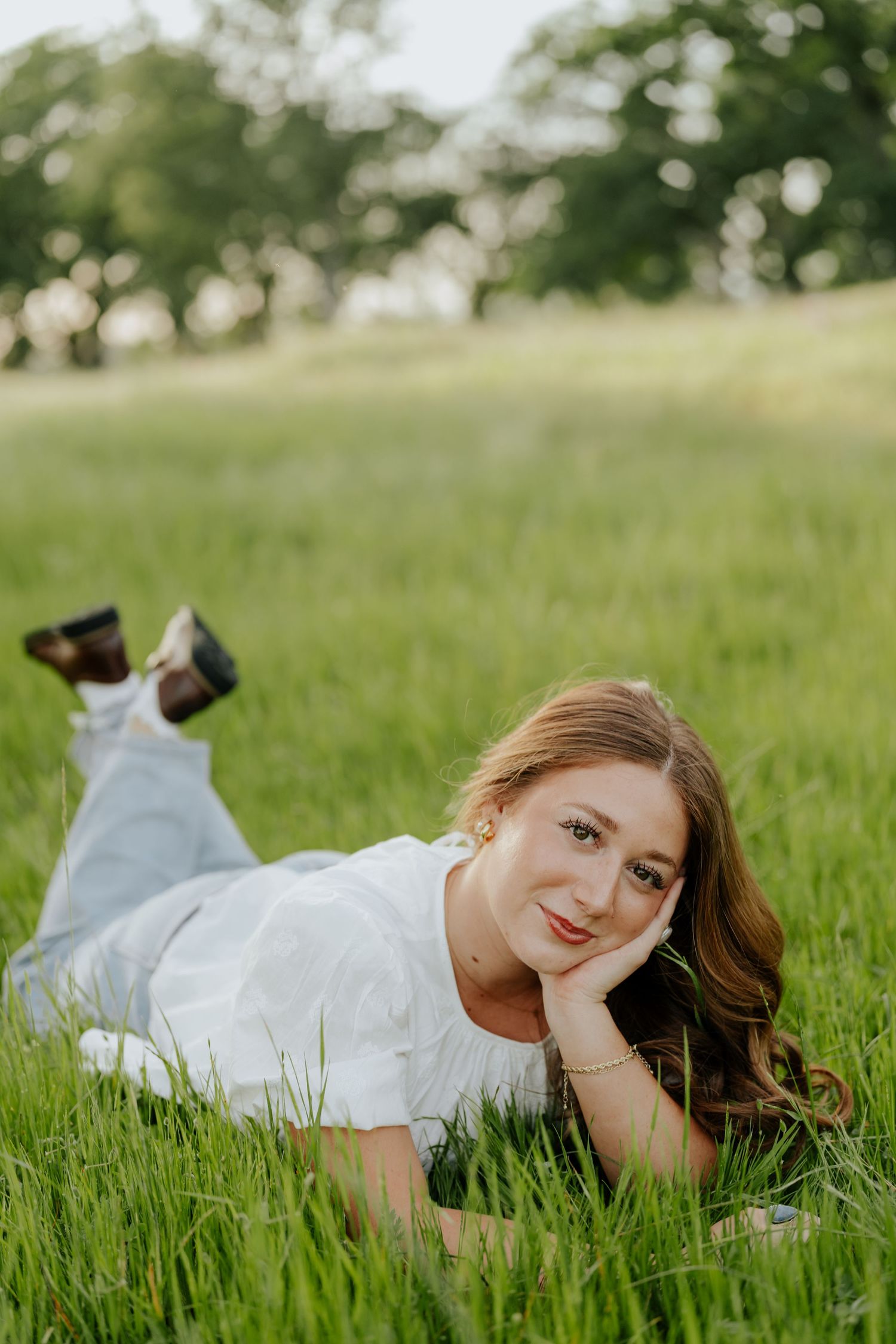 Young woman (high school senior in Sacramento, CA) in white top and jeans lying in green grass field smiling at camera.