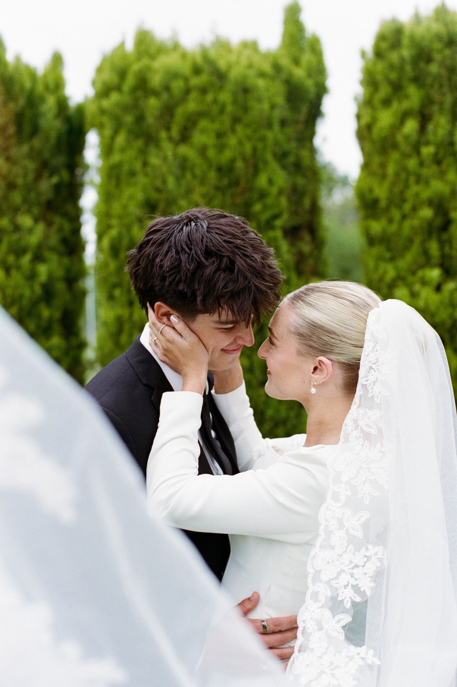 Bride and groom share an intimate moment outdoors, with lush green trees in the background.