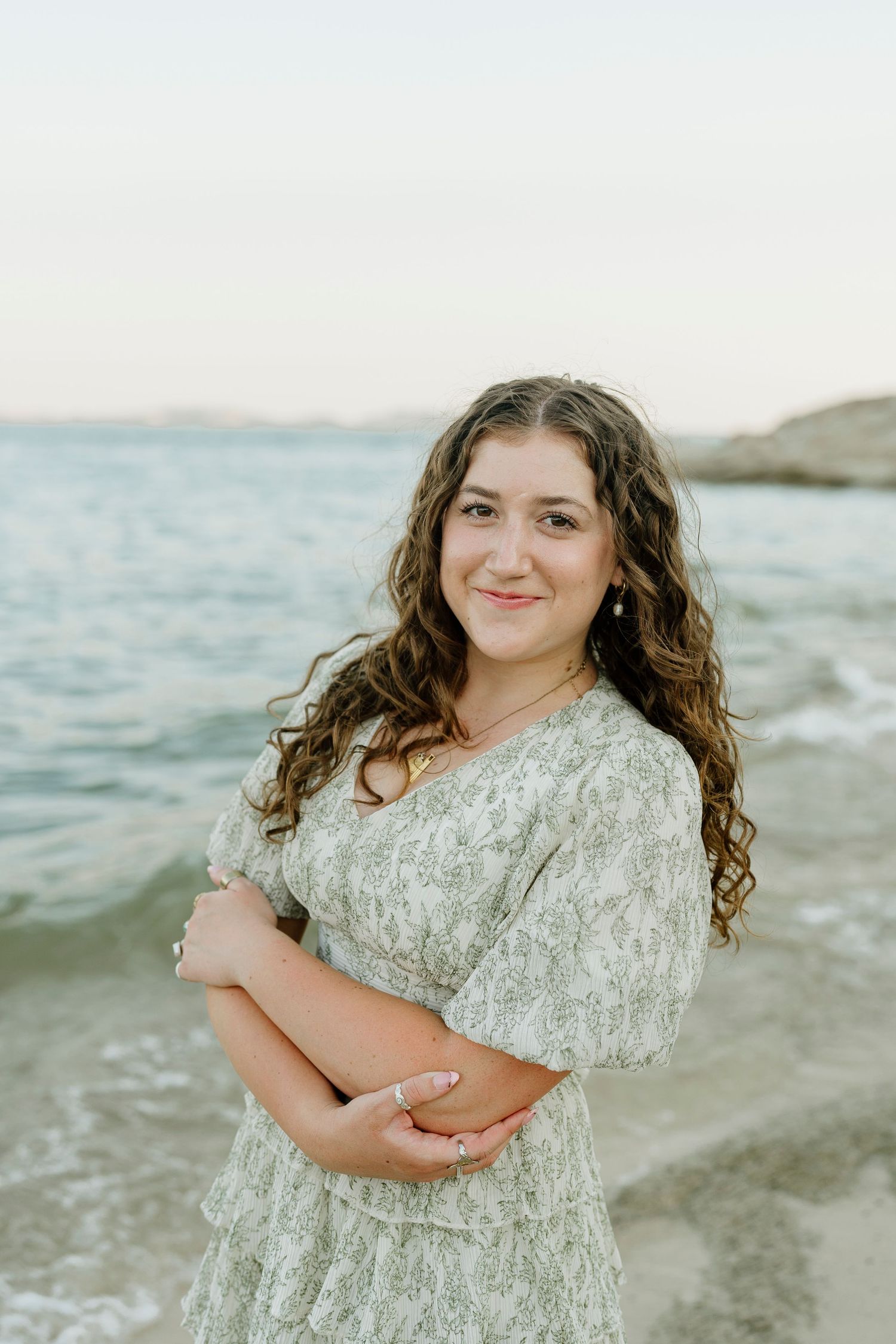 Smiling young woman in sage green floral dress stands with arms crossed at Folsom Lake at sunset.