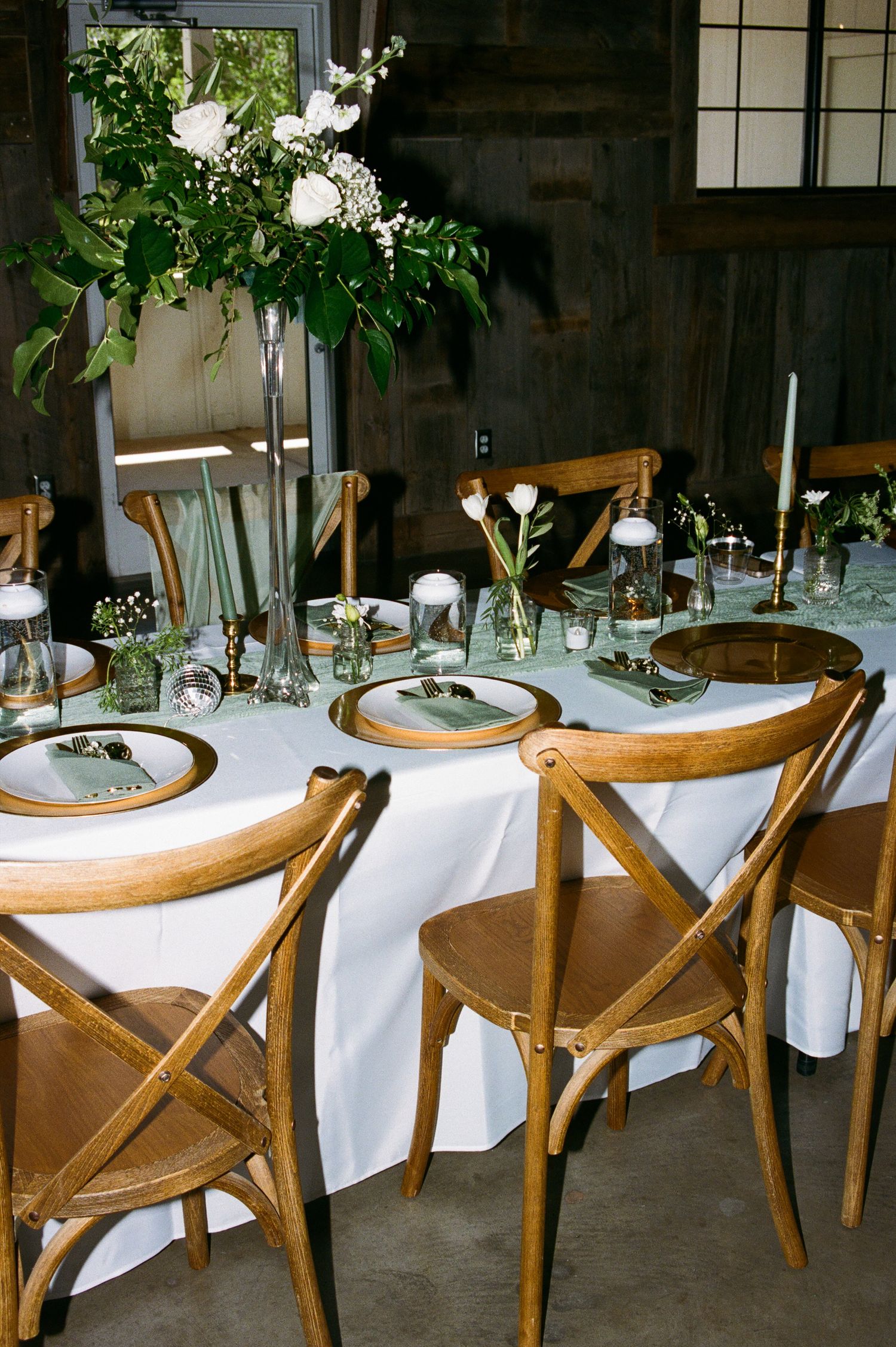 Elegant wedding reception table with white linens, wooden cross-back chairs, and tall greenery centerpiece.