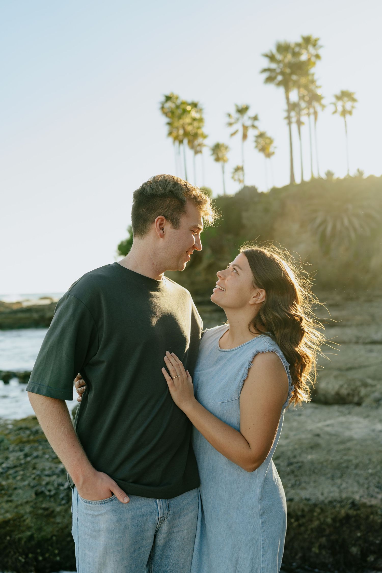 Couple gazing at each other smiling on rocky beach with palm trees at sunset in Laguna Beach, California.