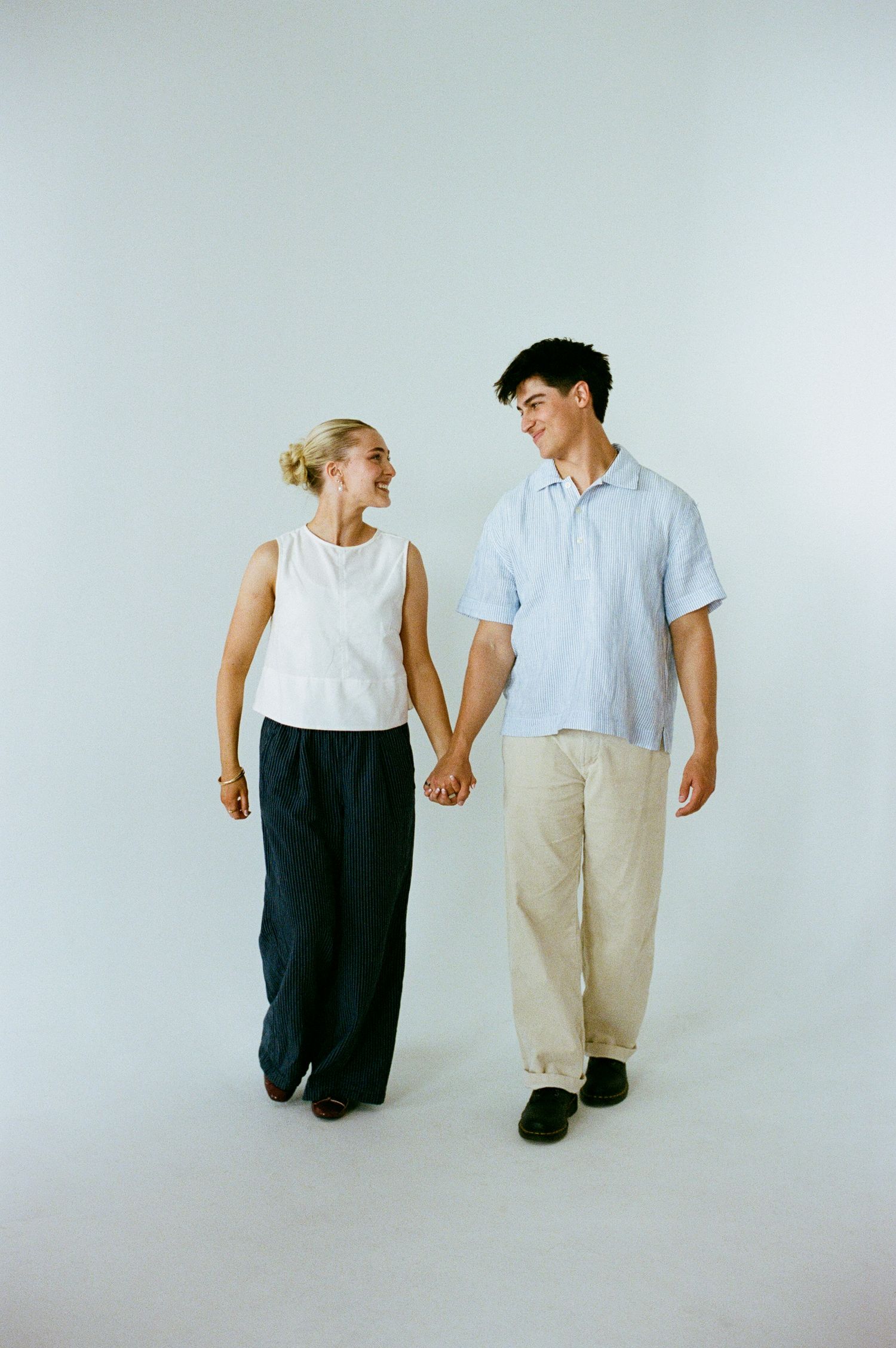 Young couple holds hands and smiles at each other against a white studio background.