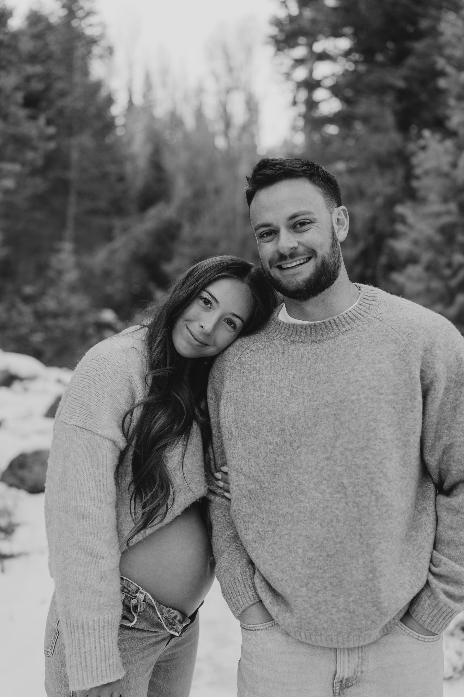 Black and white photo of smiling couple during maternity shoot in snowy forest setting.