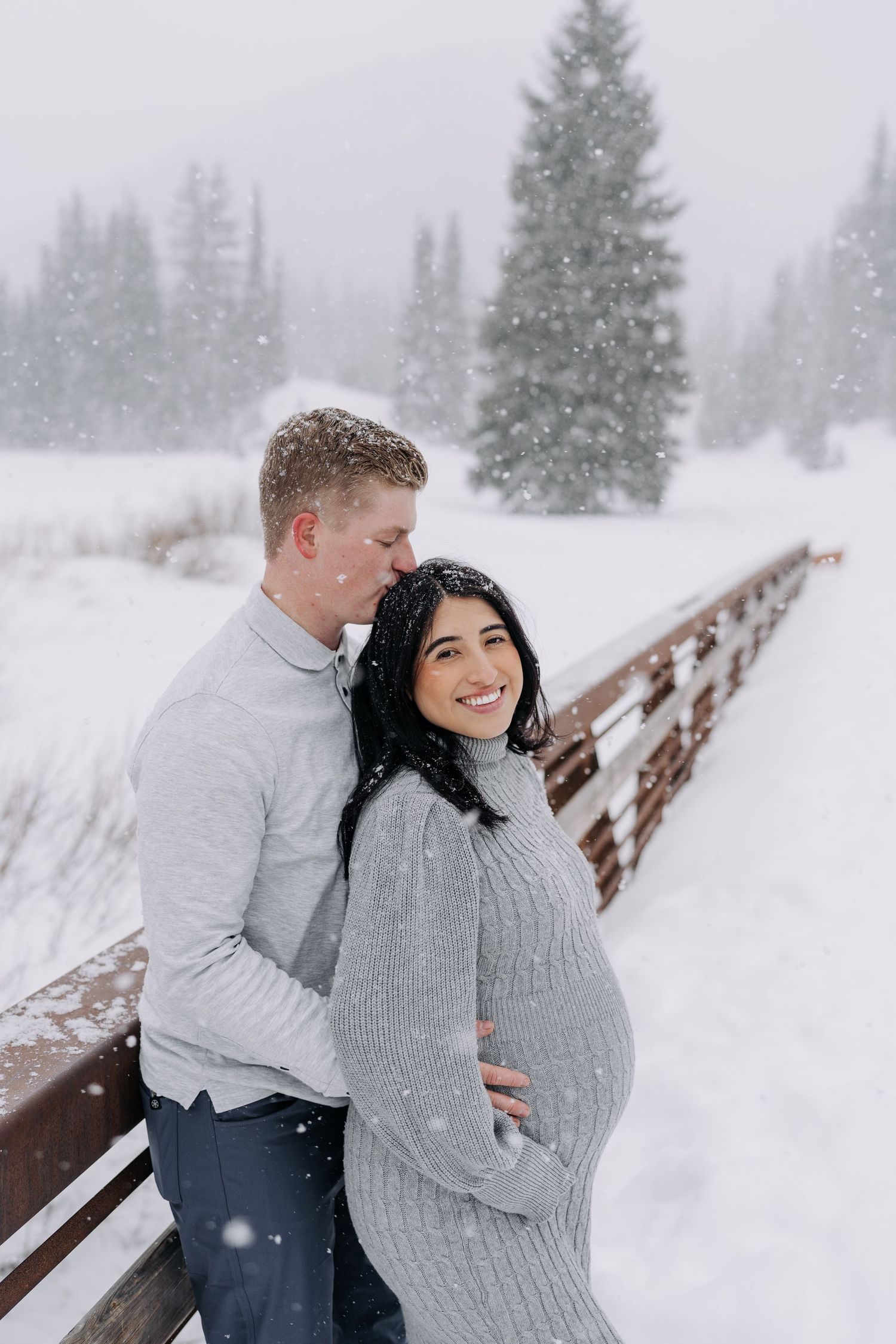 Smiling pregnant woman in grey knit dress embraced by partner on snowy bridge during snowfall.
