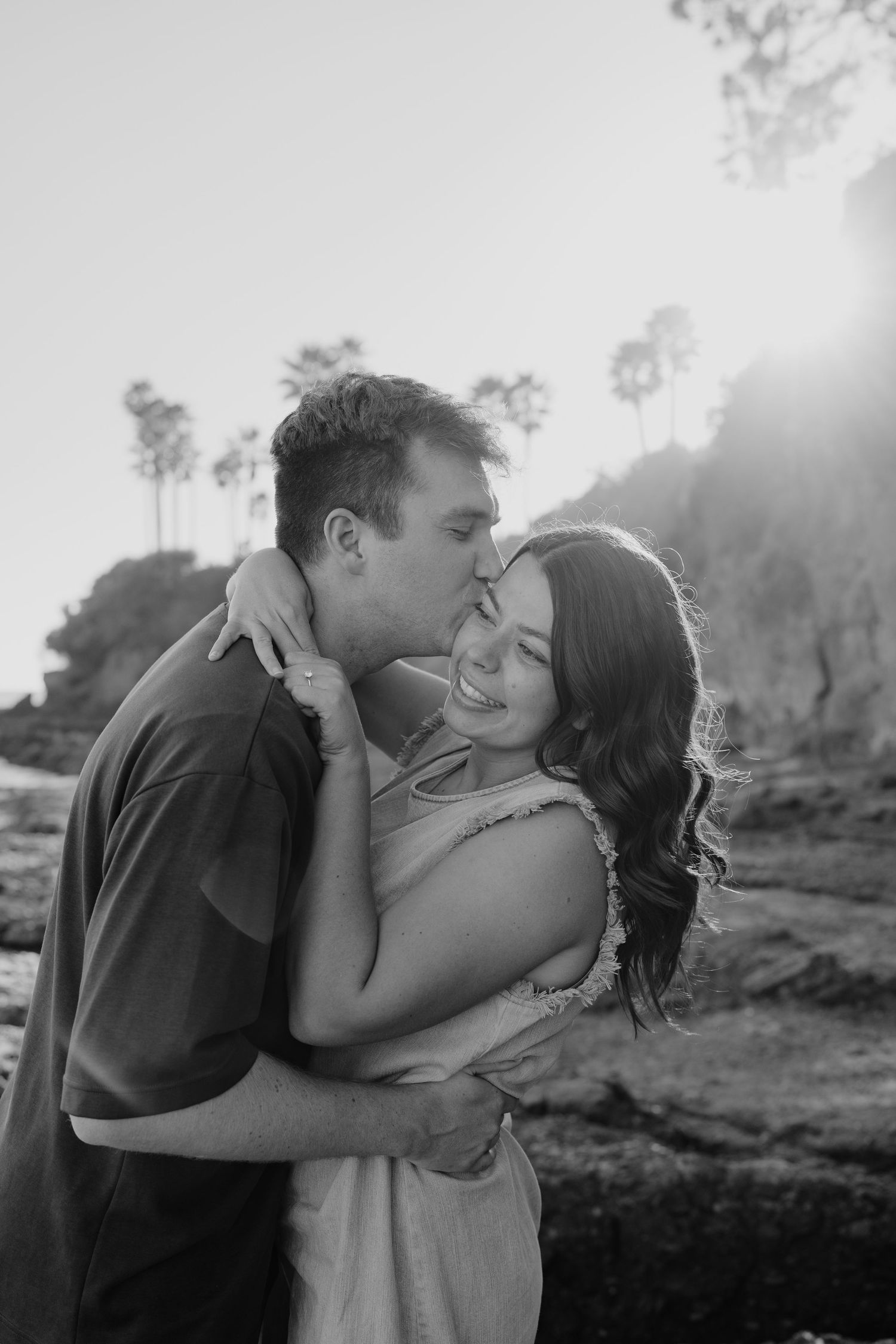 Black and white photo of man kissing smiling woman's cheek near rocky coastal cliffs in Laguna Beach, California.