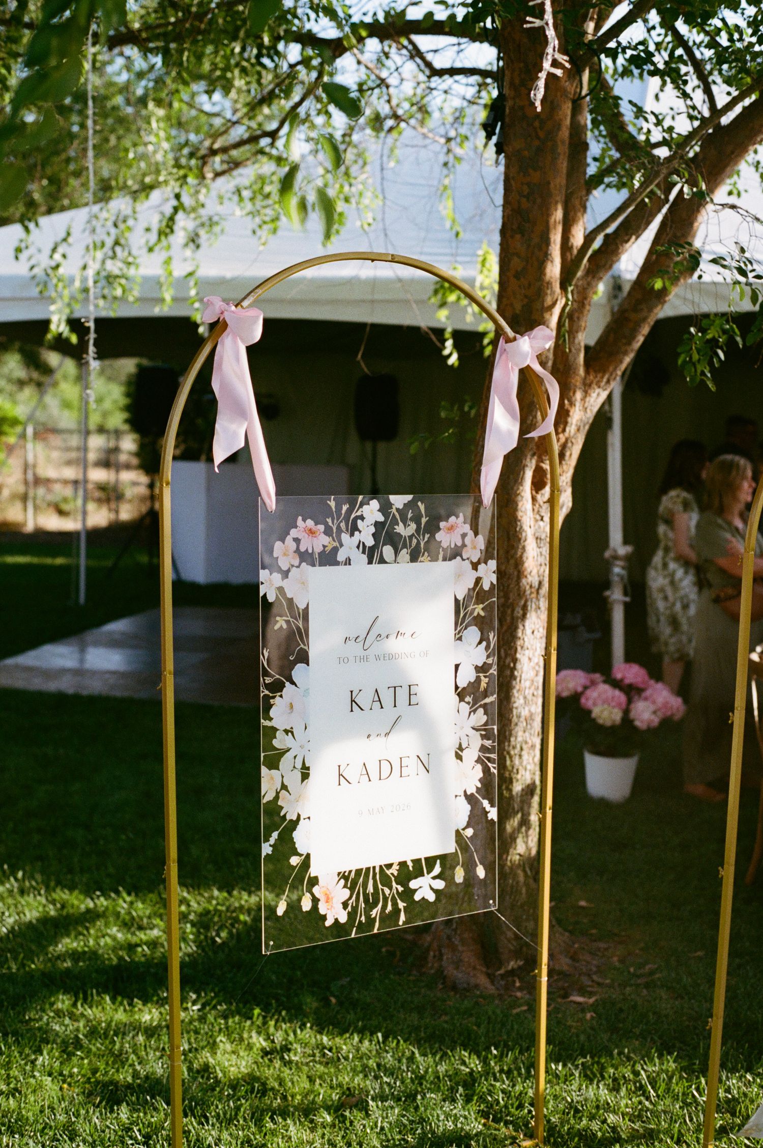 Elegant wedding welcome sign for Kate and Kaden, hung with pink ribbons on a gold arch stand outdoors.