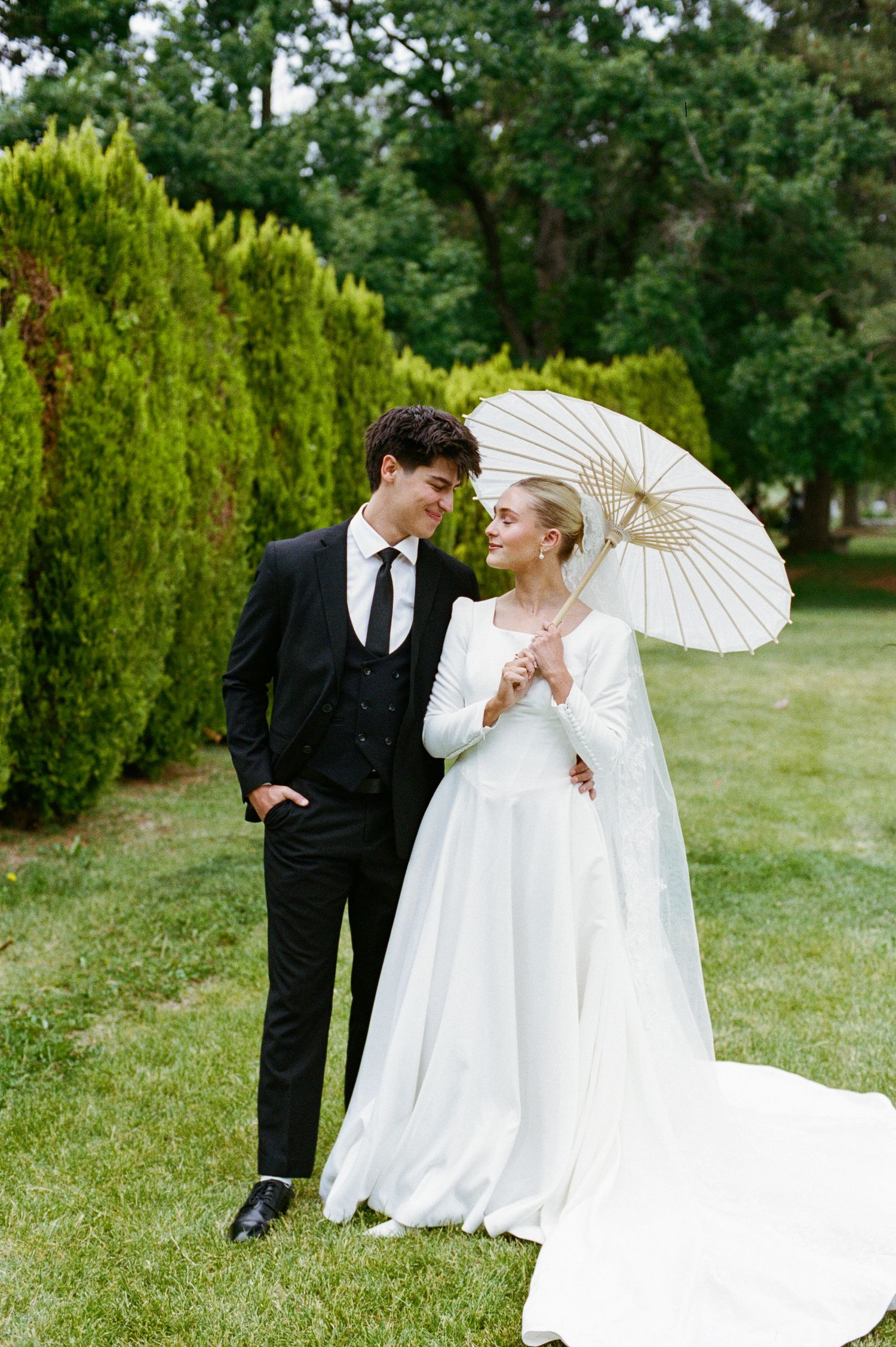 A bride in a white gown holding a parasol stands with a groom in a black suit in a lush green garden.