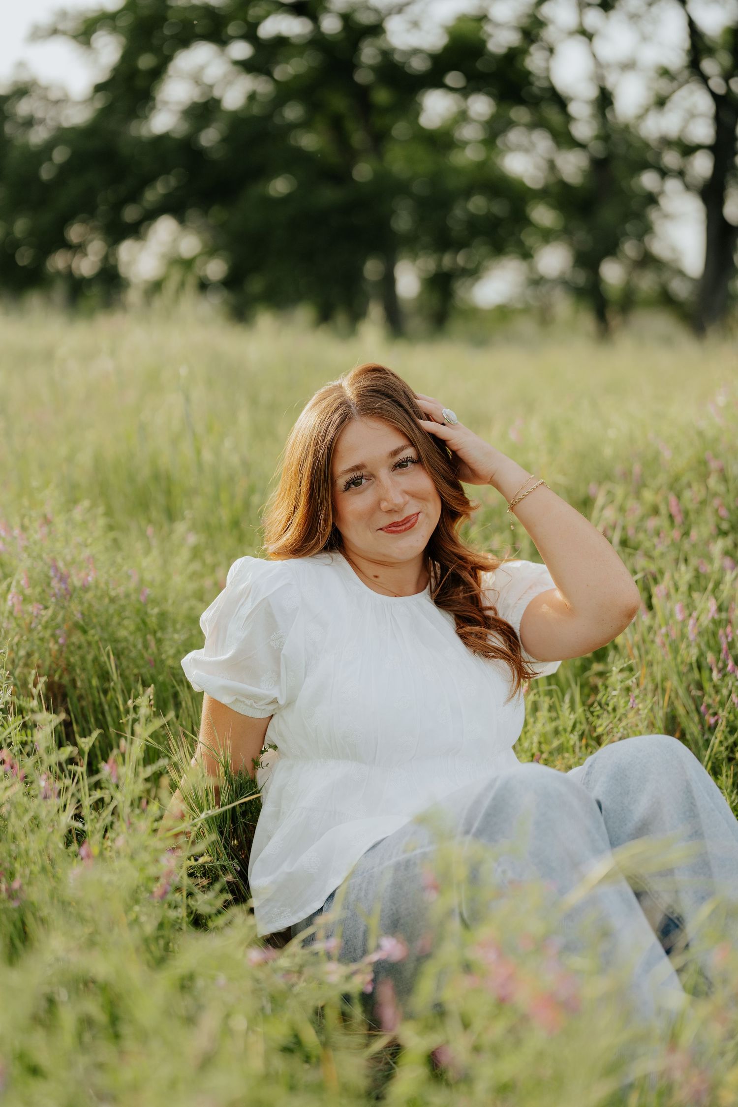 Smiling redhead woman (high school senior in Sacramento, CA) in white blouse sitting in wildflower meadow during golden hour.