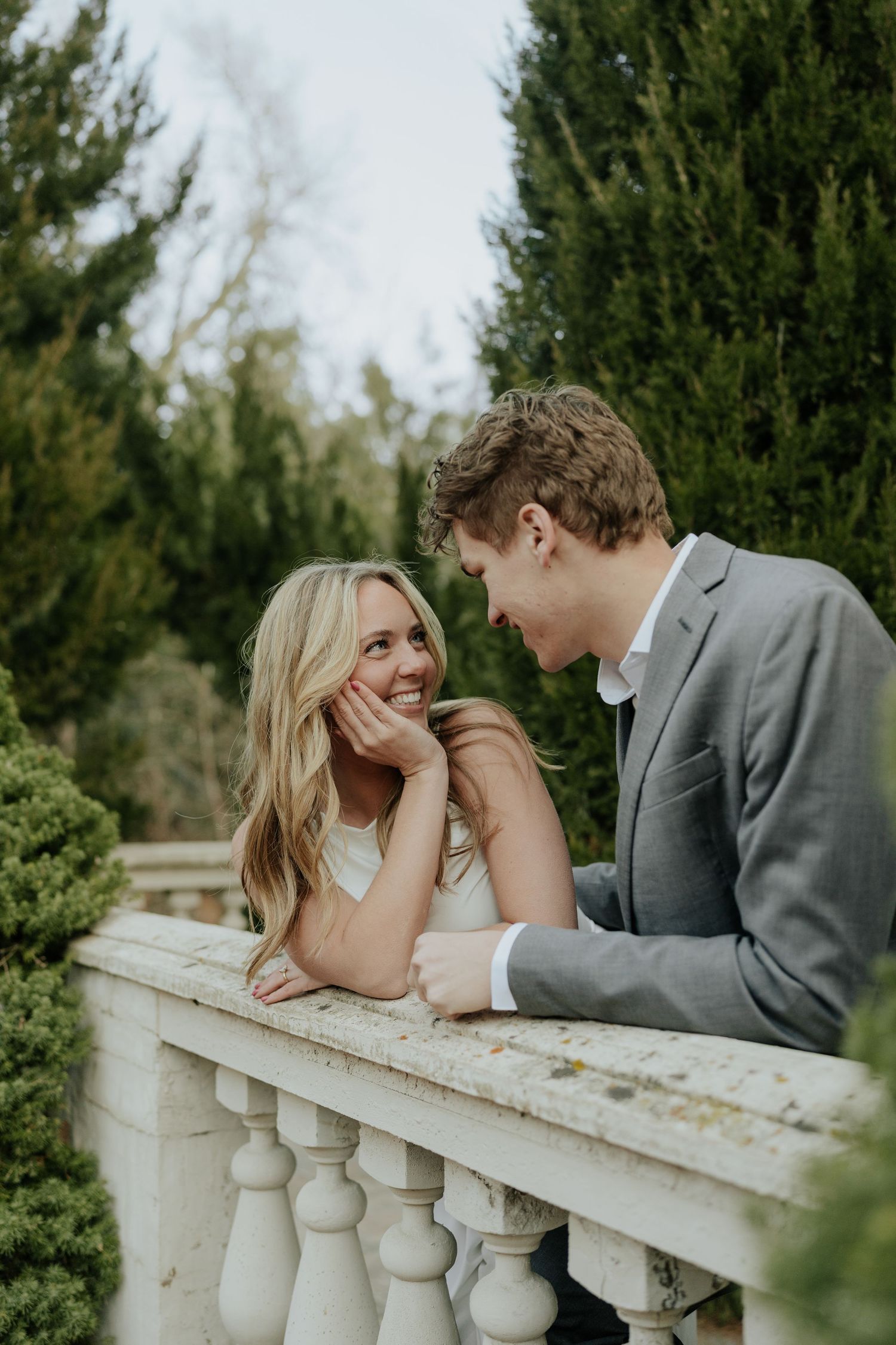 Couple smiling at each other leaning on a stone balustrade surrounded by tall green cypress trees at La Caille Restaurant in Utah.