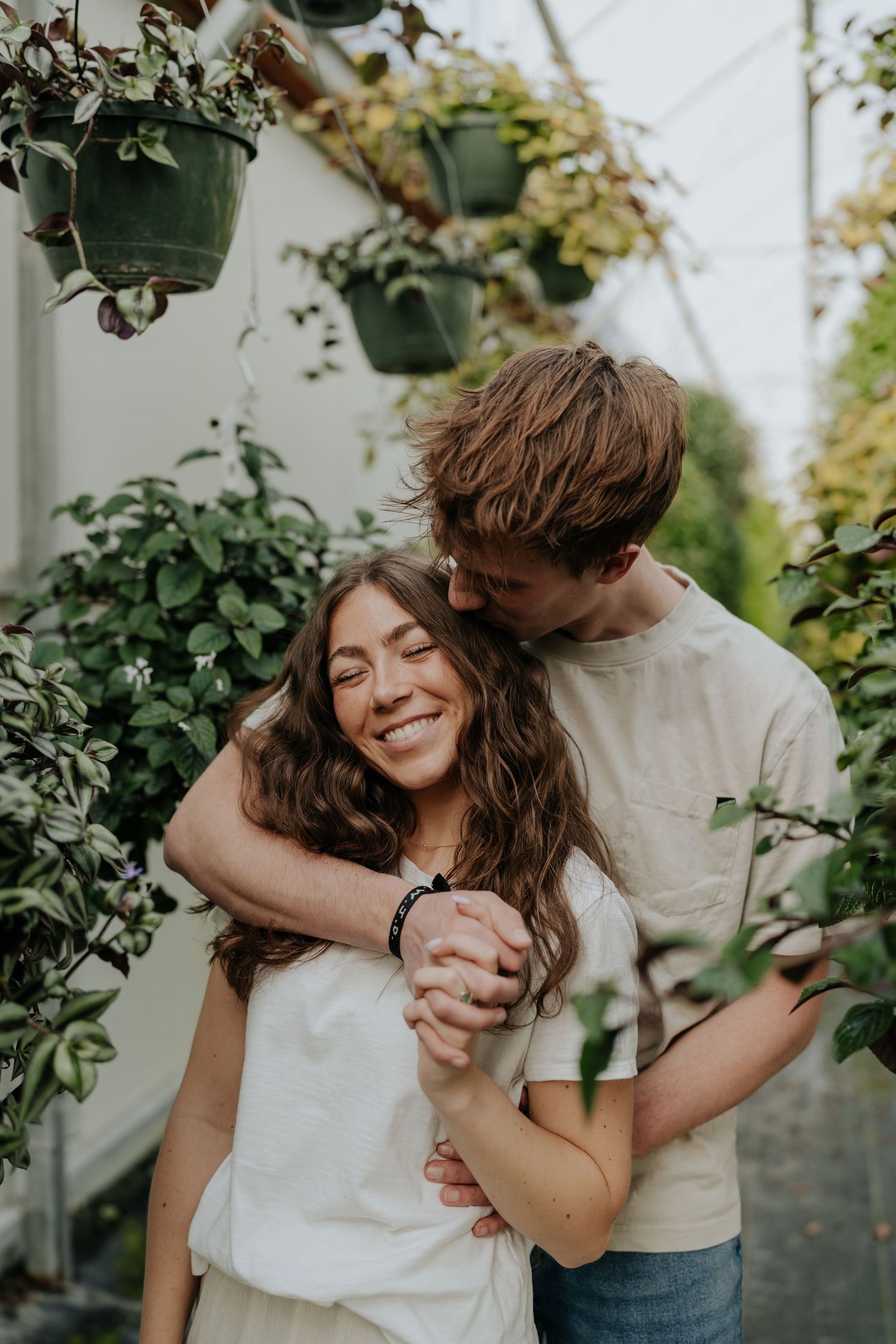 Man kissing smiling woman on the cheek in a plant-filled greenhouse during engagement photos in Provo, Utah.