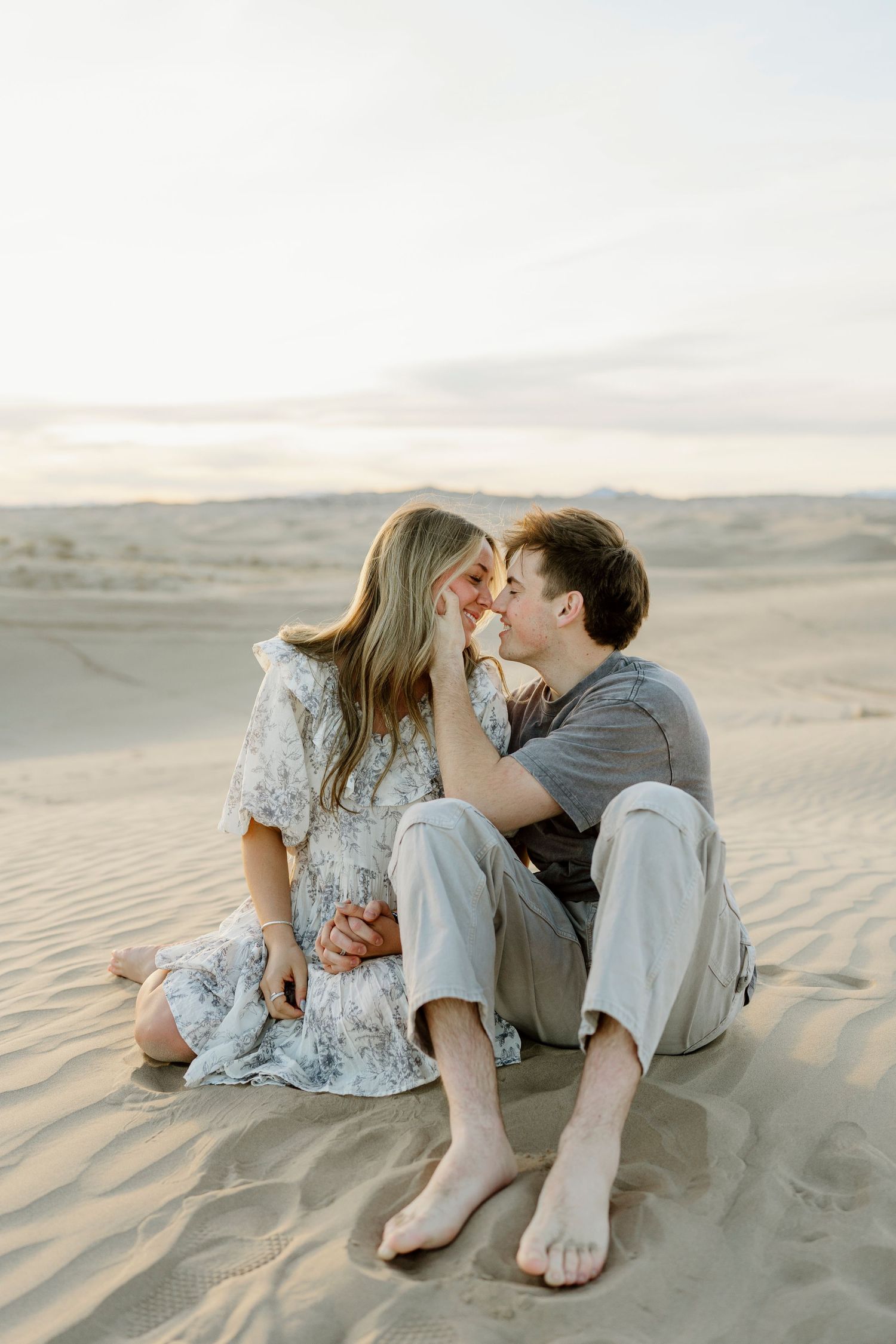 Couple sits intimately on sand dunes sharing a tender moment during desert engagement session.
