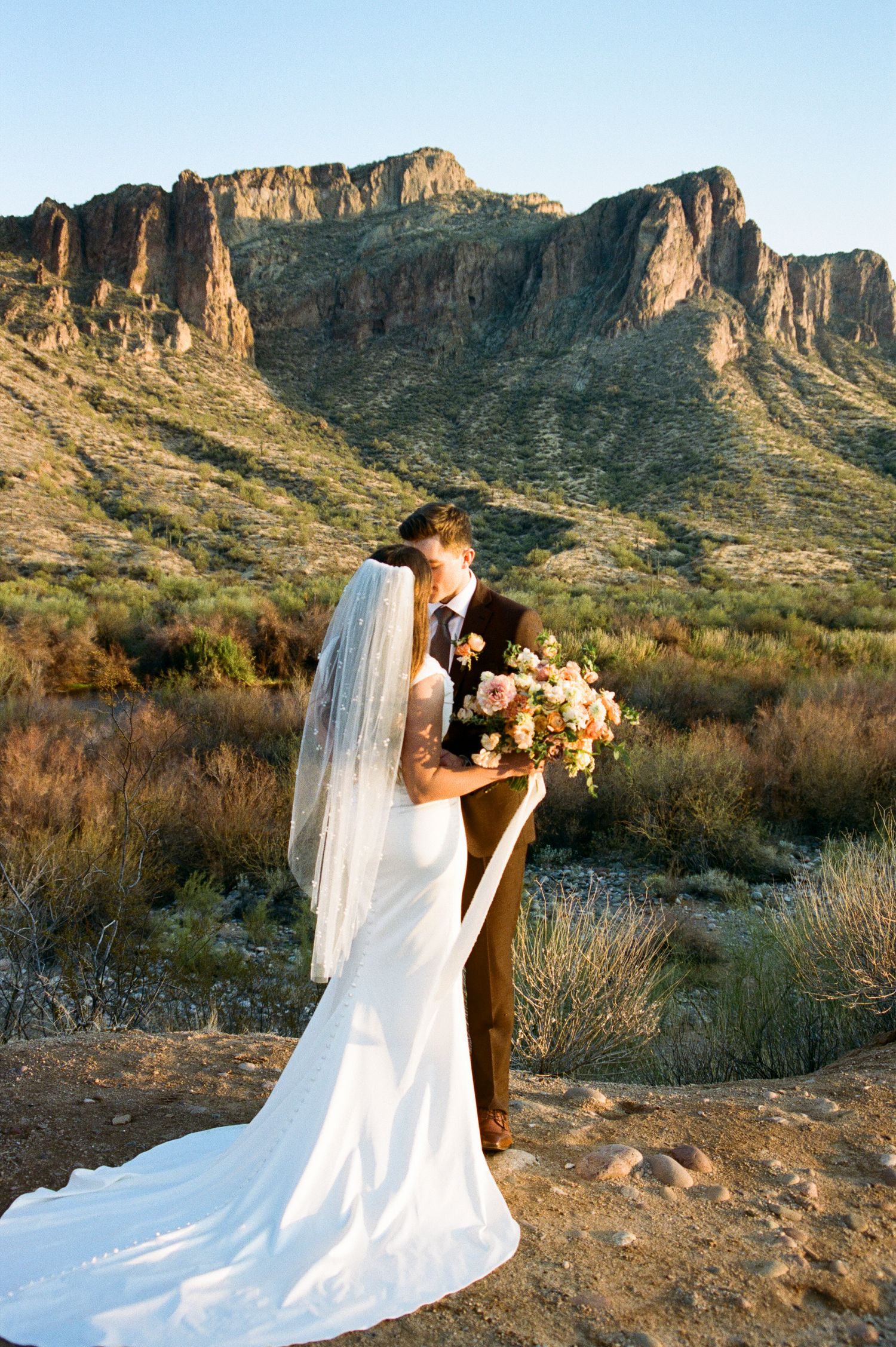 Bride and groom share a romantic kiss in the desert with stunning mountain backdrop at sunset.