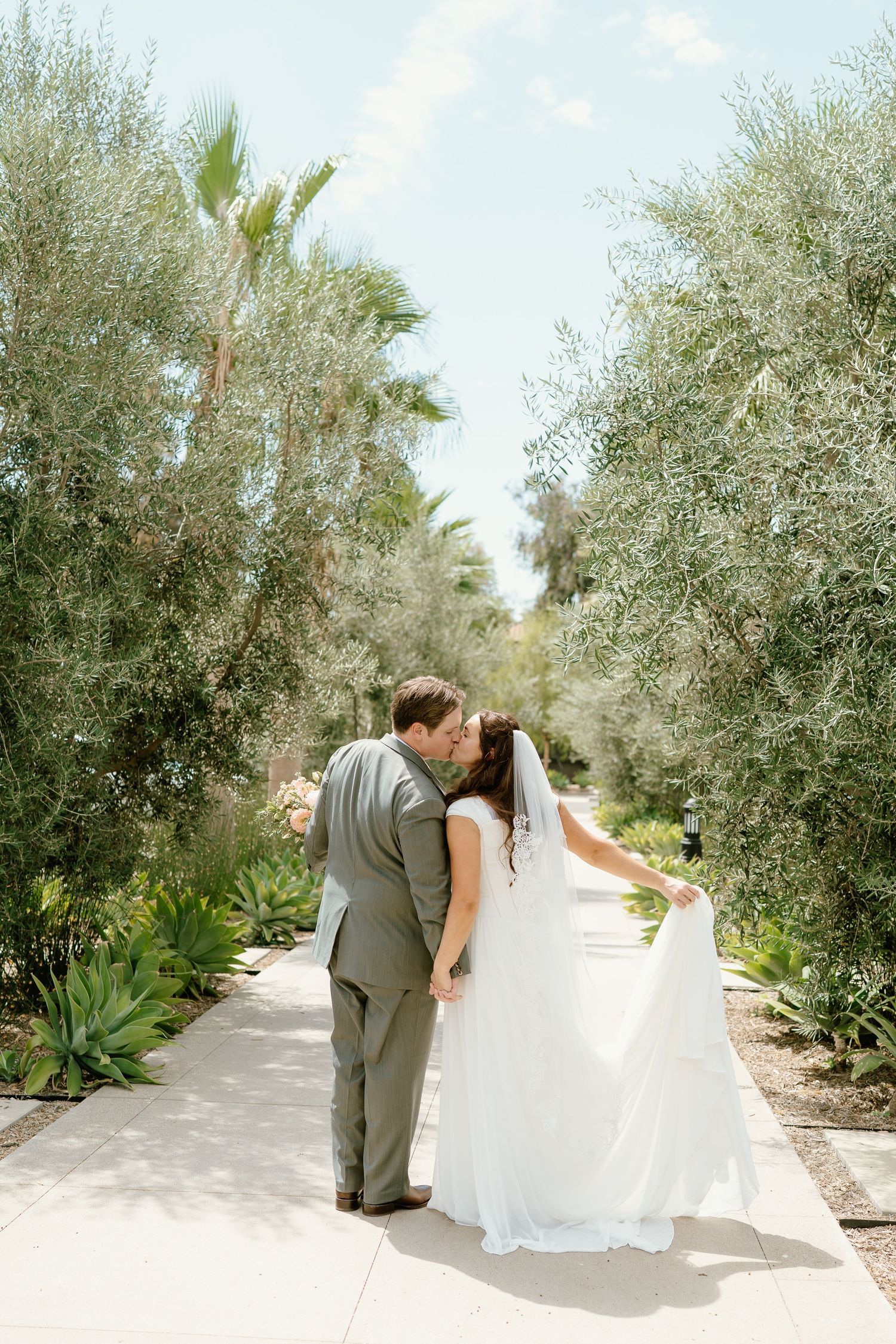 Bride and groom share a romantic kiss on a garden path lined with olive and palm trees on their wedding day.