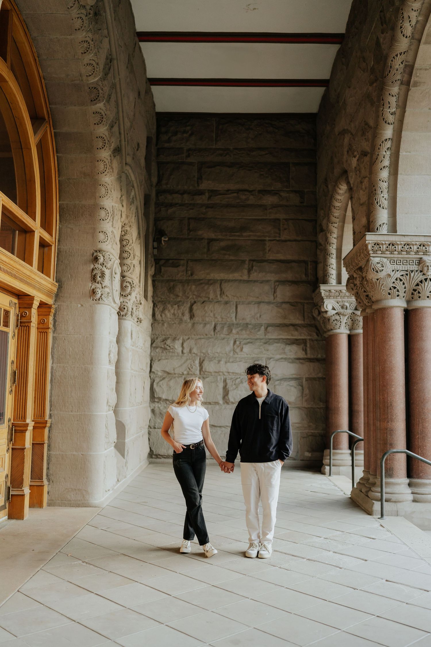 Couple holding hands walking through ornate stone archway corridor during engagement photo session.