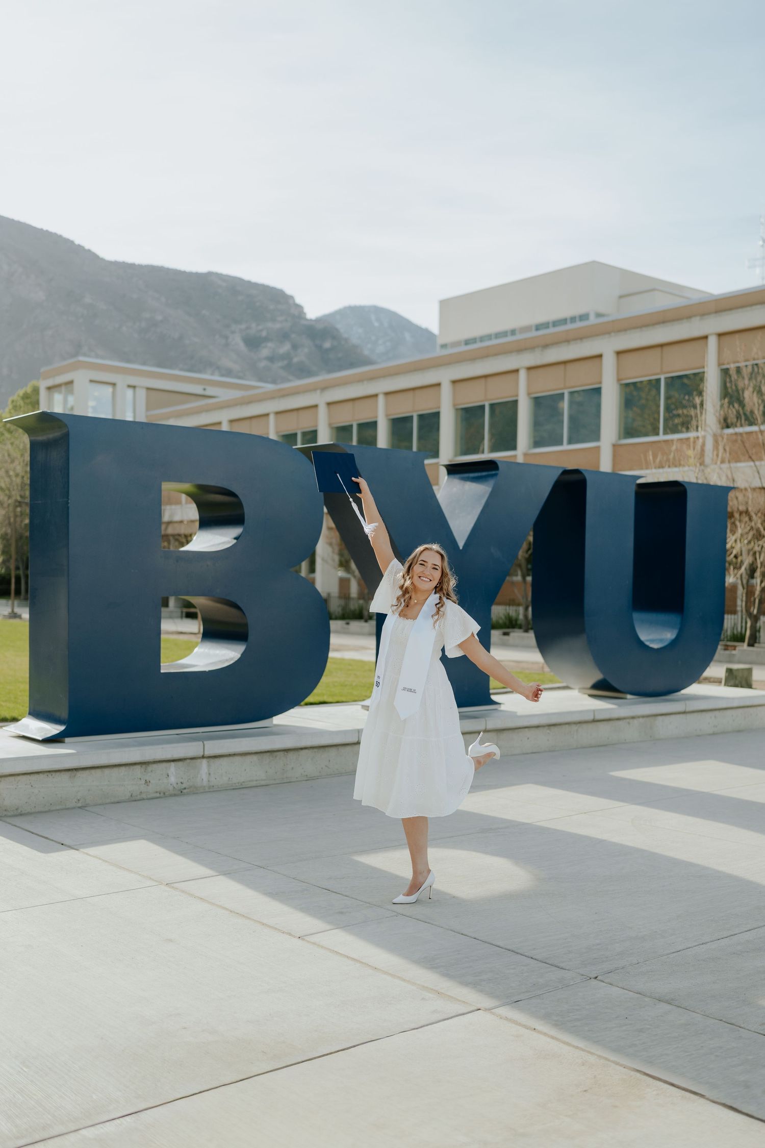 Woman in white dress celebrating graduation in front of large BYU letters sign on Brigham Young University campus.