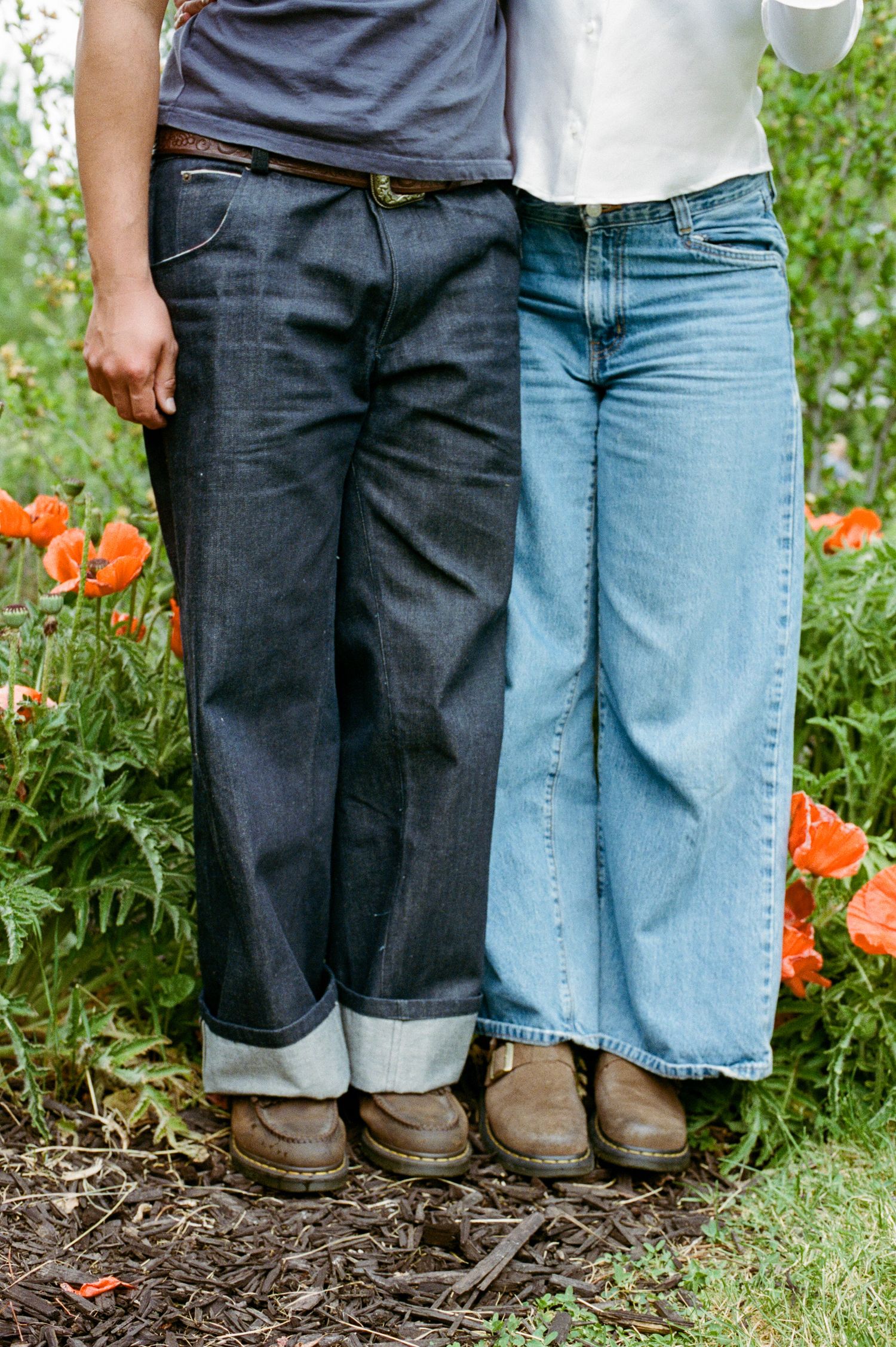 Two people wearing wide-leg jeans and brown boots standing in a garden with red poppy flowers.
