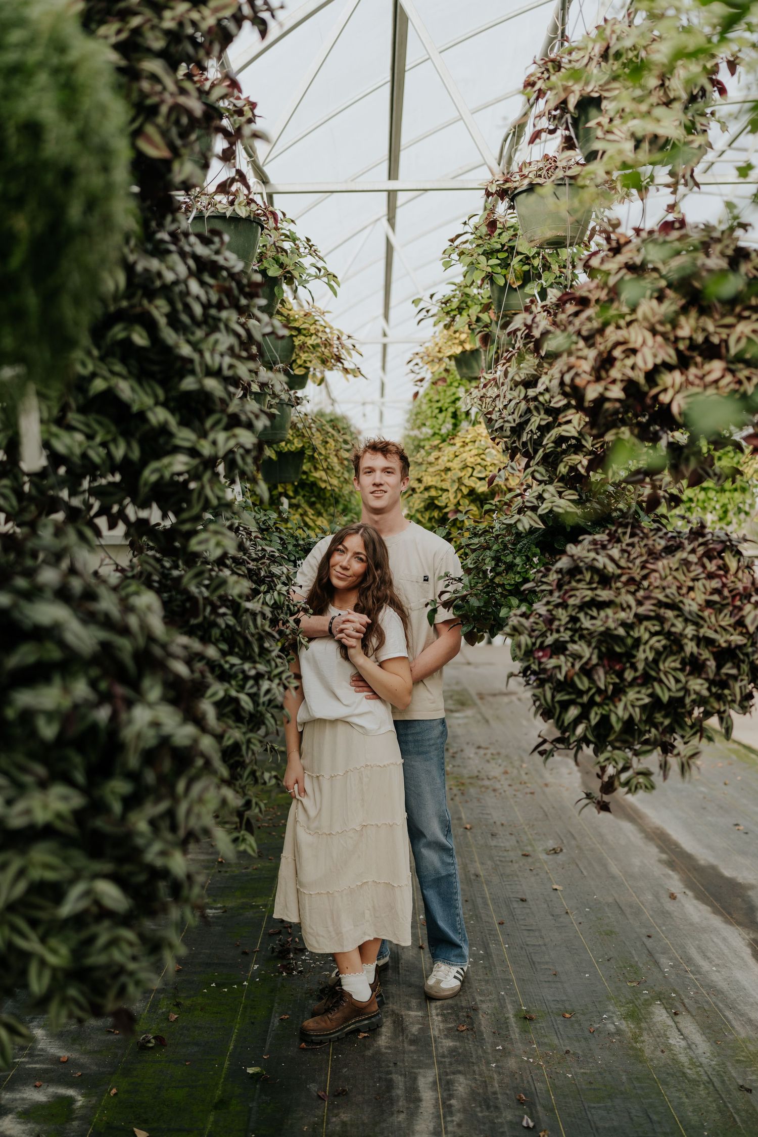 Couple embracing in a greenhouse aisle surrounded by hanging plants and lush green foliage in Provo, Utah.