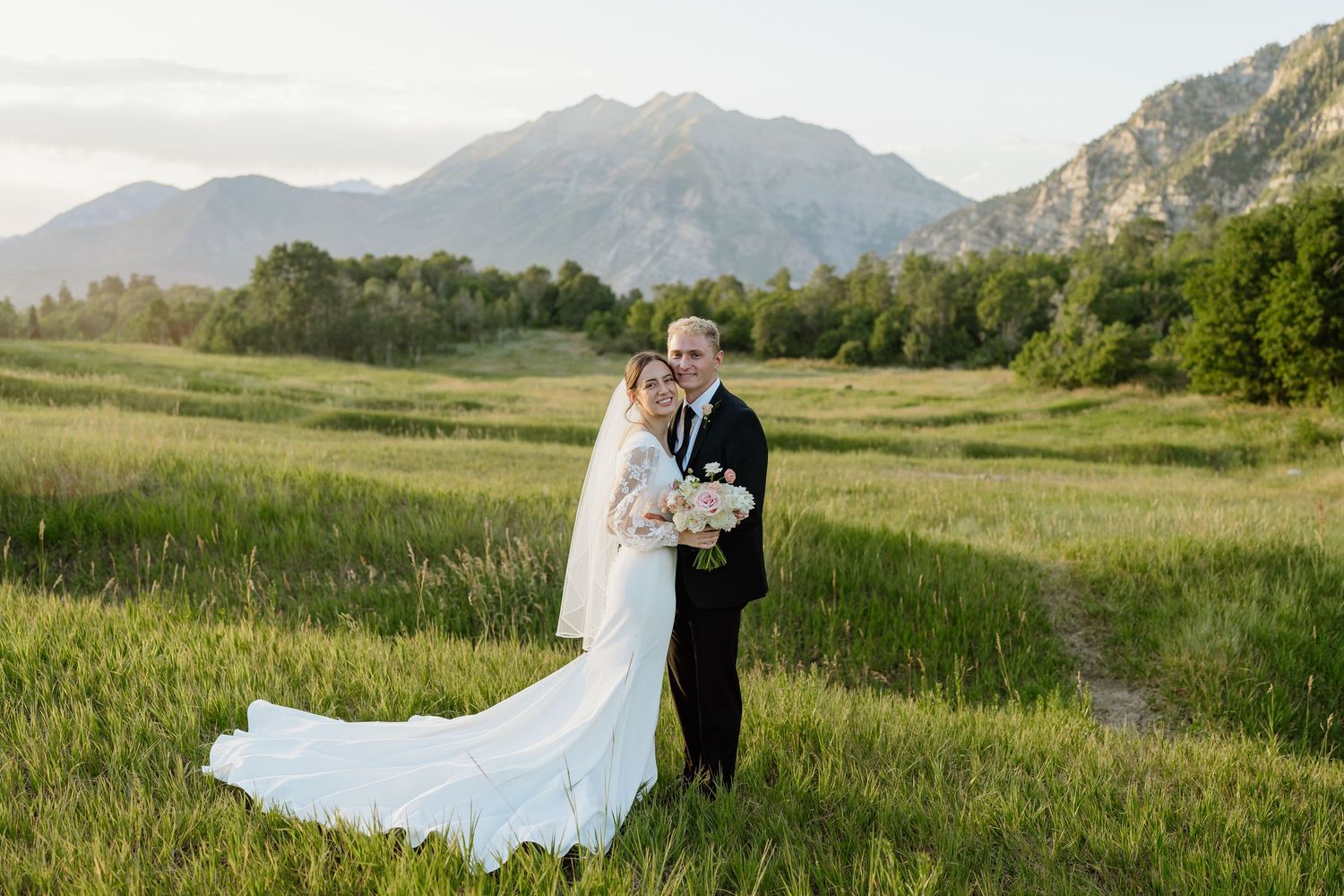 Bride and groom posing in green field with mountain range and clear sky backdrop near Provo, Utah.