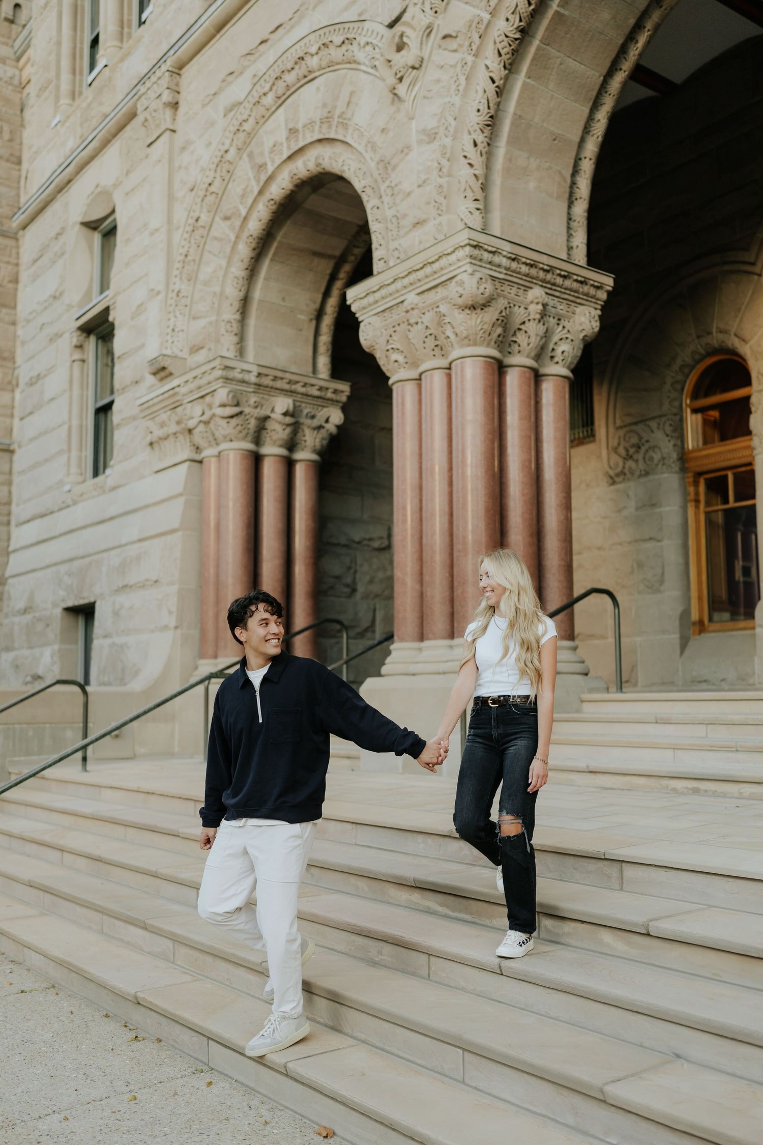 Couple holding hands descending steps of ornate historic stone building with grand columns.