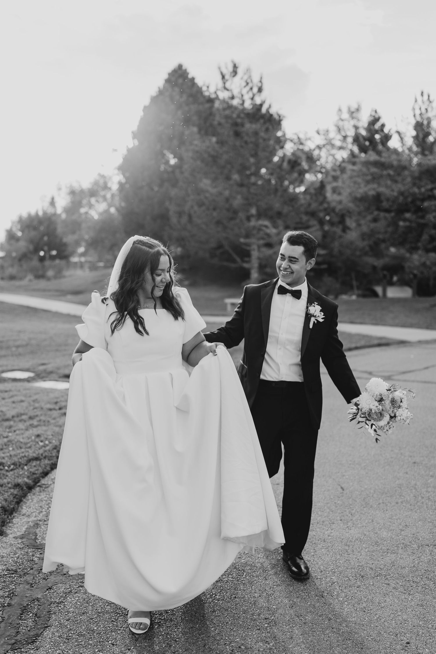 Black and white photo of bride and groom walking and laughing together down a sunlit path.
