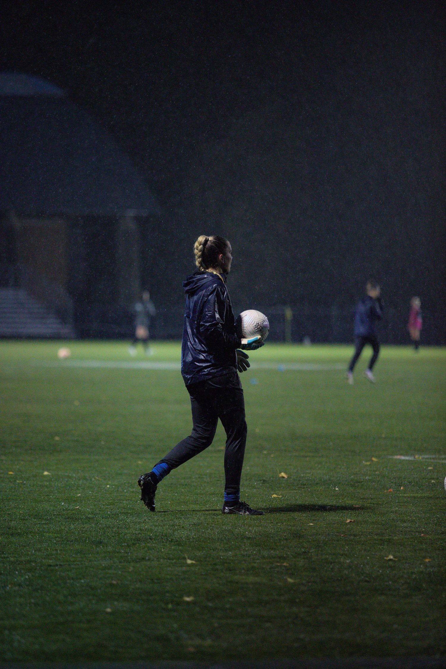 A goalkeeper holds a soccer ball on a rain-soaked field under bright lights during a night training session.