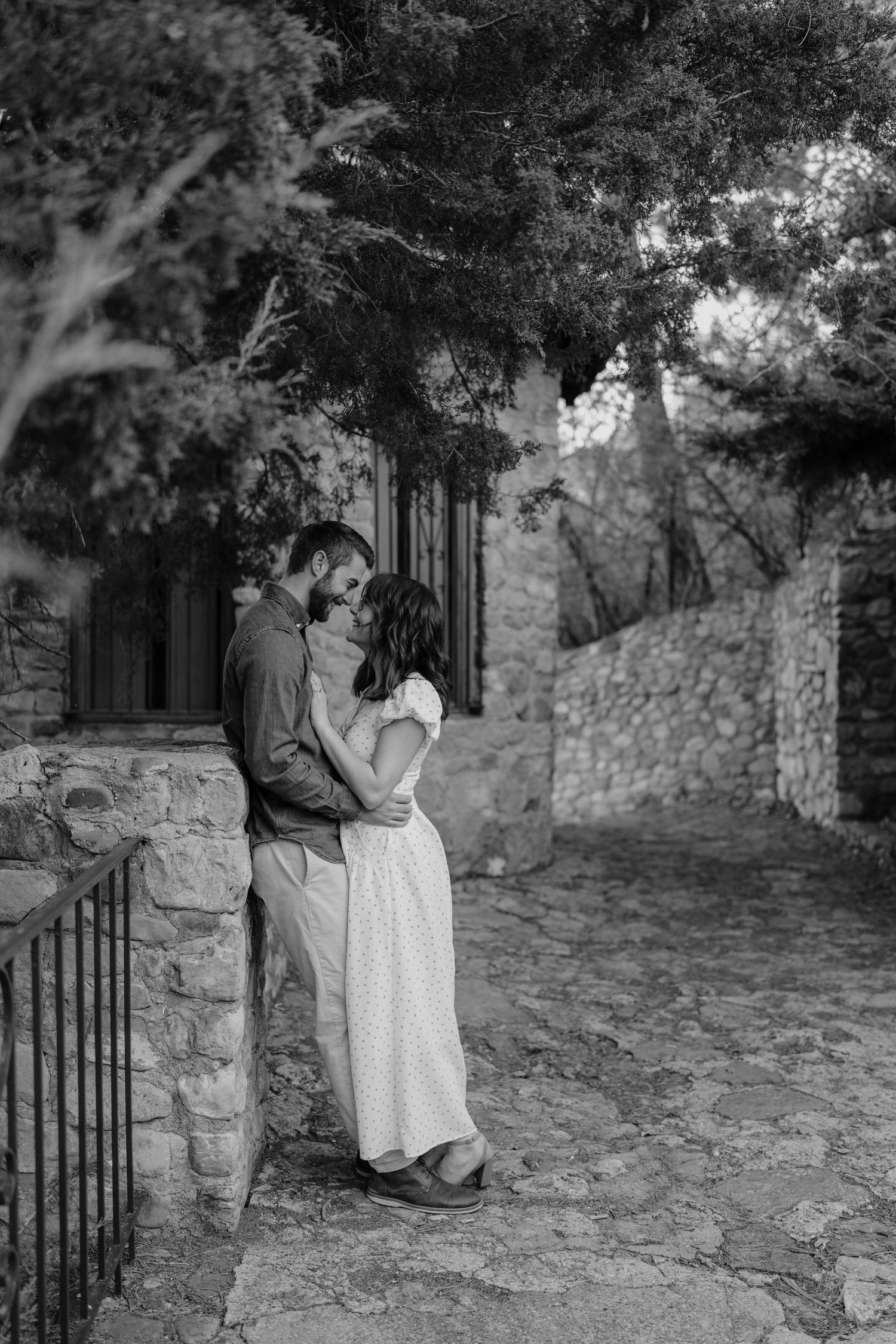 Black and white photo of couple embracing tenderly against a rustic stone wall outdoors.