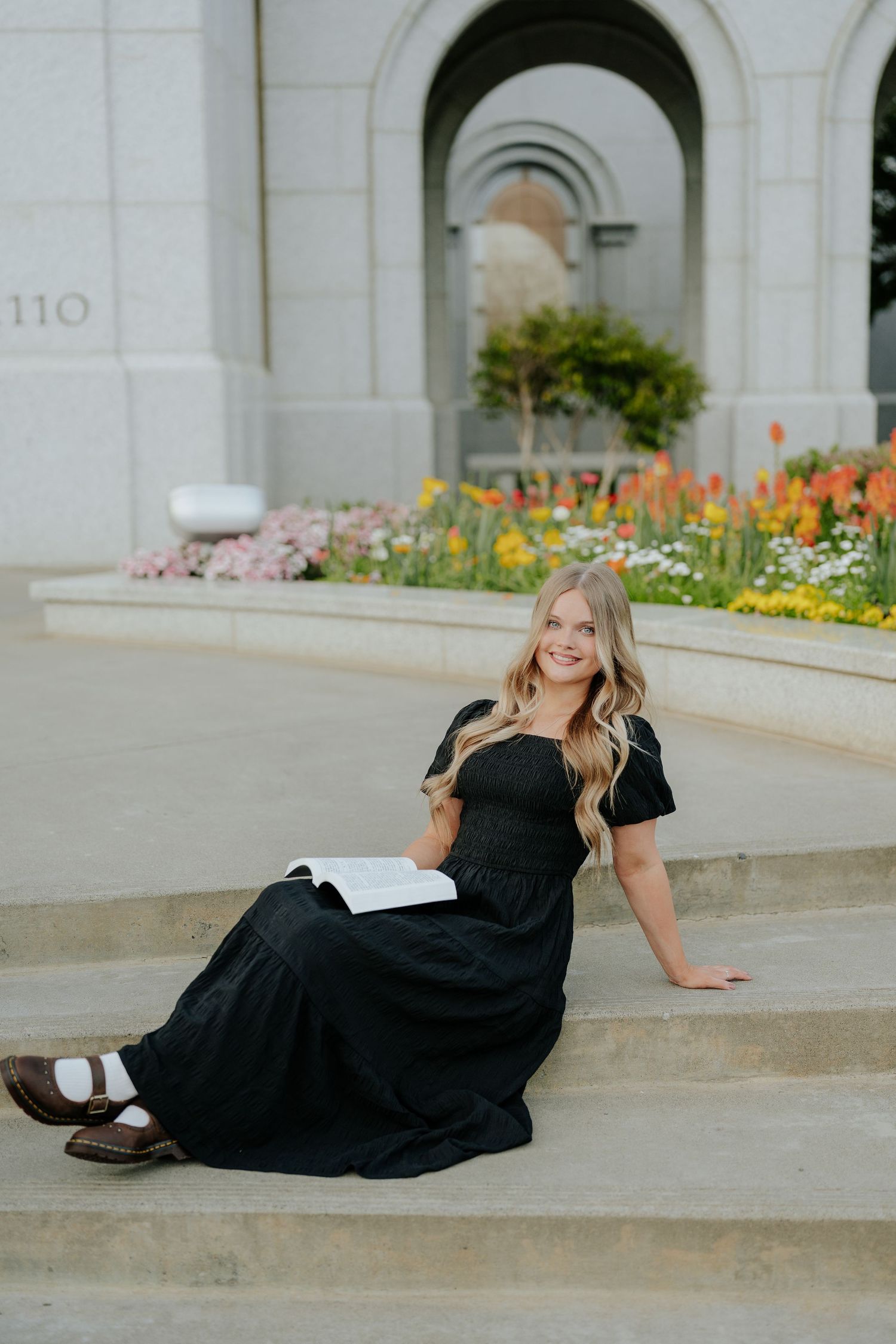 Smiling blonde woman in black dress sitting on steps with open book near flower garden at Sacramento LDS temple.