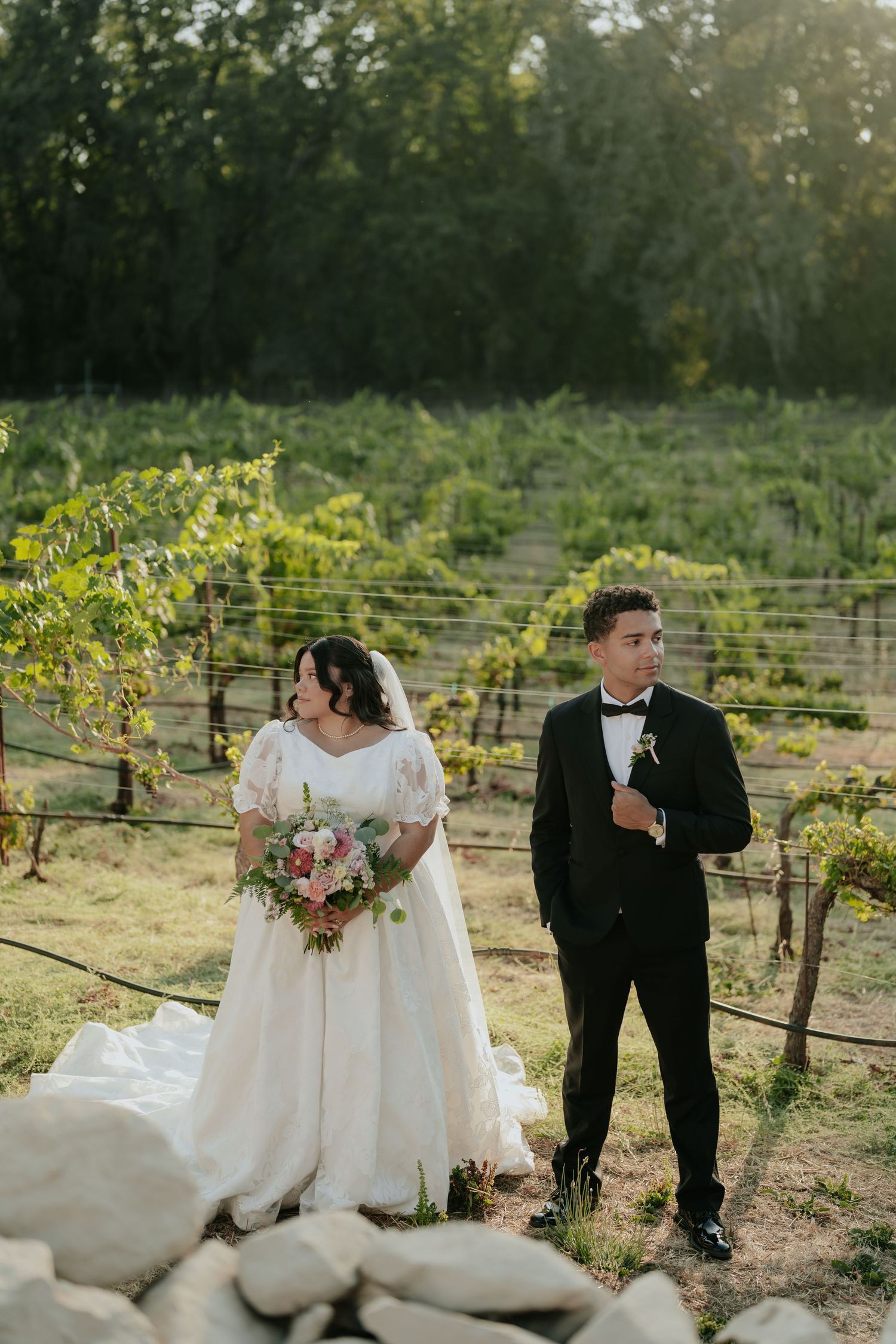 Bride and groom posing in a vineyard, bride holding a floral bouquet in a white gown, groom in a black tuxedo.