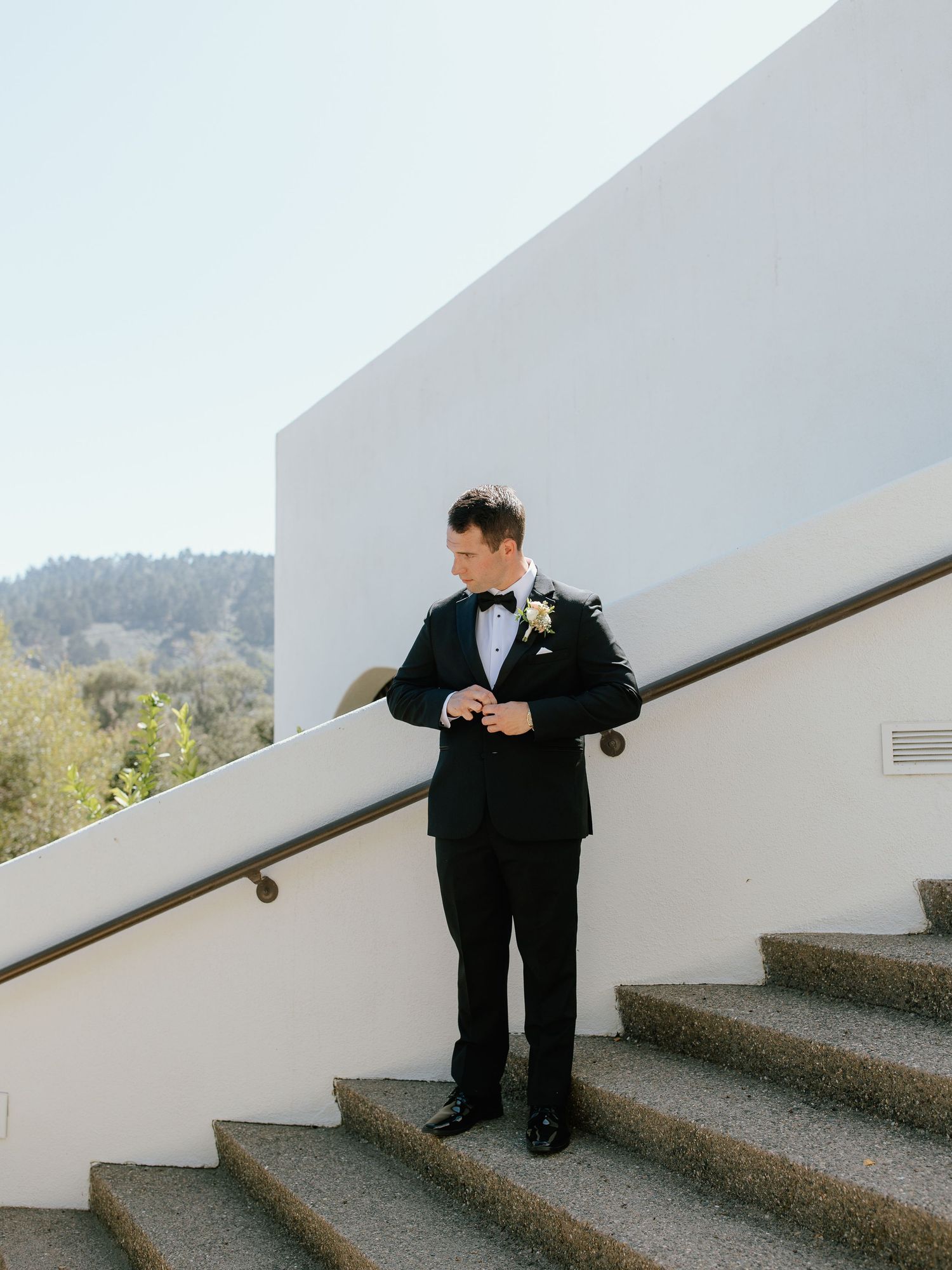 Groom in black tuxedo adjusting cufflinks on modern white staircase with scenic hillside backdrop.