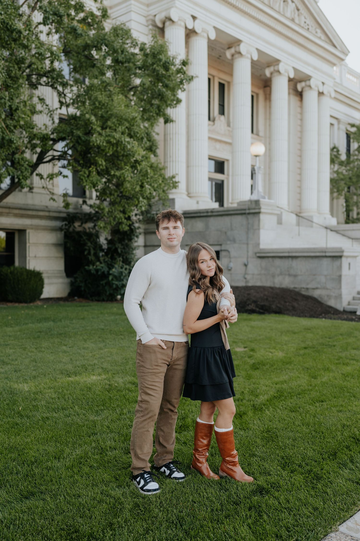 Young couple stands together on green lawn before white neoclassical building, woman in black dress and boots.