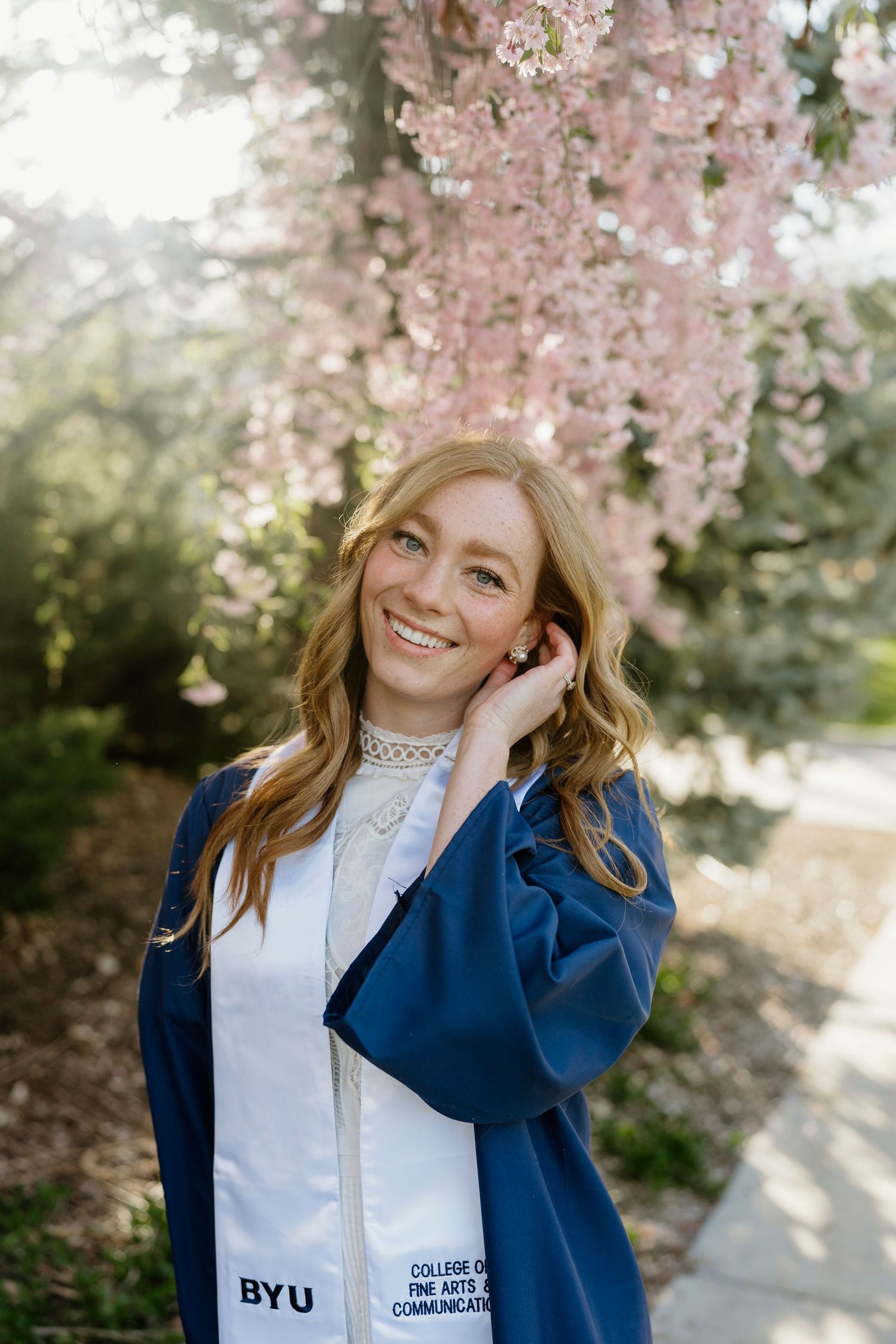 Smiling BYU graduate in blue gown with Fine Arts and Communications stole under pink blossoms.