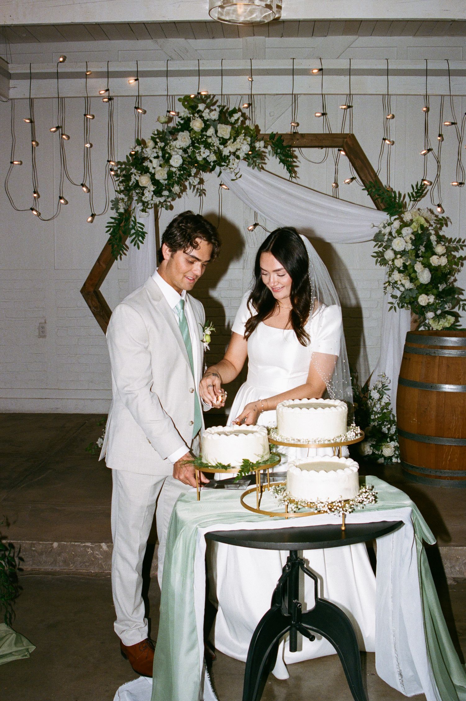 Newlyweds cut a three-tier white wedding cake beneath a hexagonal arch with string lights and white florals.