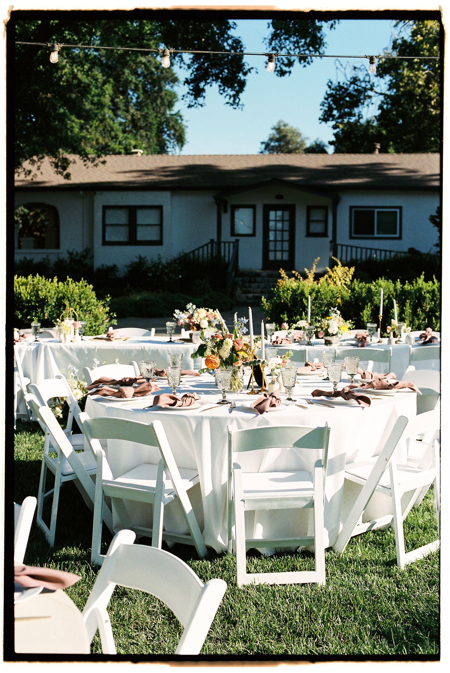 Elegant outdoor wedding reception with round tables, white chairs, floral centerpieces, and string lights on green lawn.