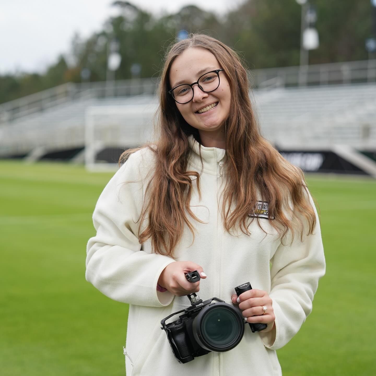 A smiling young woman with long hair holds a professional camera on a soccer field.