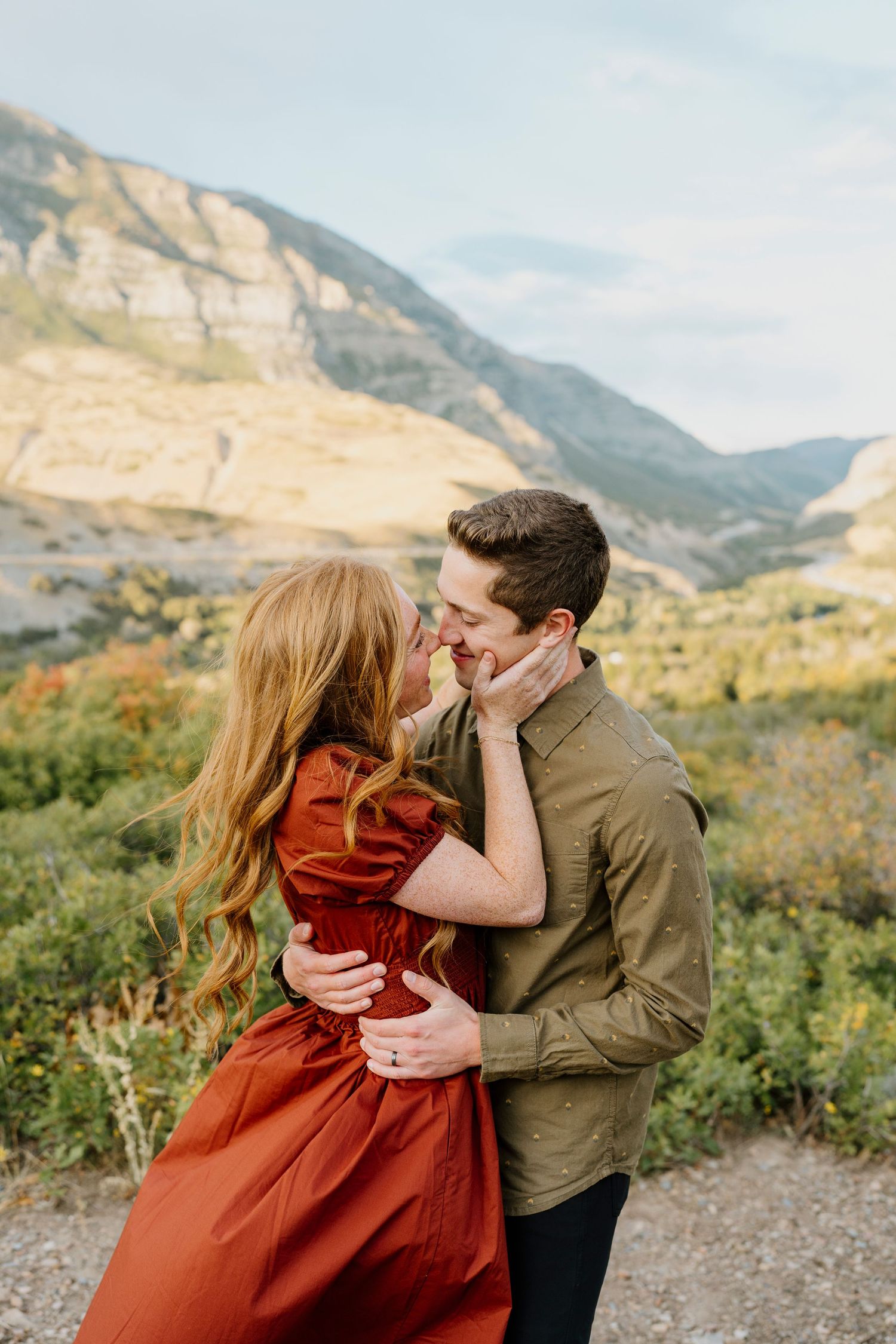 Couple nearly kissing in an autumn mountain landscape, woman in red dress and man in olive shirt.