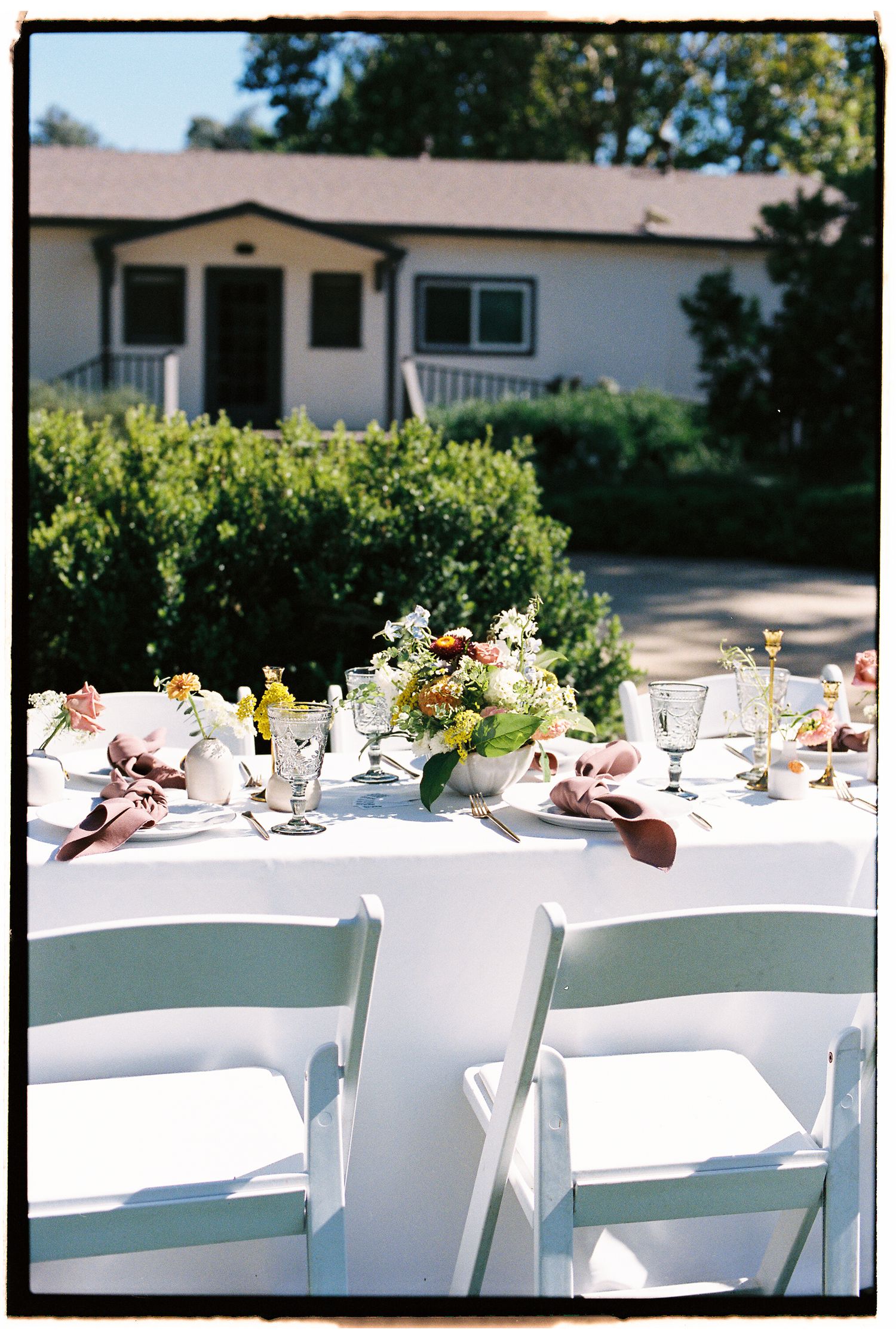 Elegant outdoor wedding reception table with white chairs, floral centerpiece, and glassware set against a charming house backdrop.