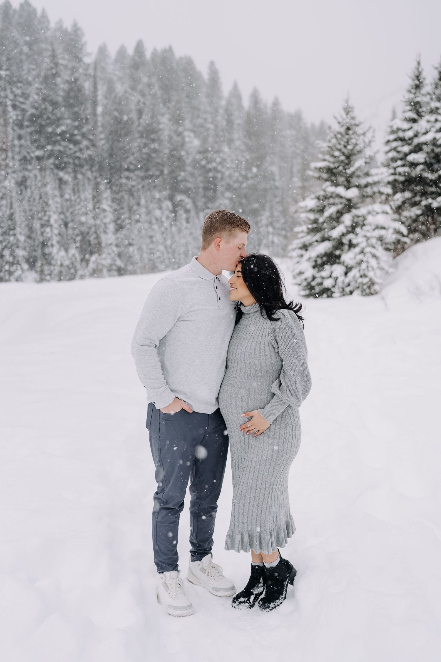 Expecting couple sharing a kiss in heavy snowfall surrounded by snow-covered pine trees.