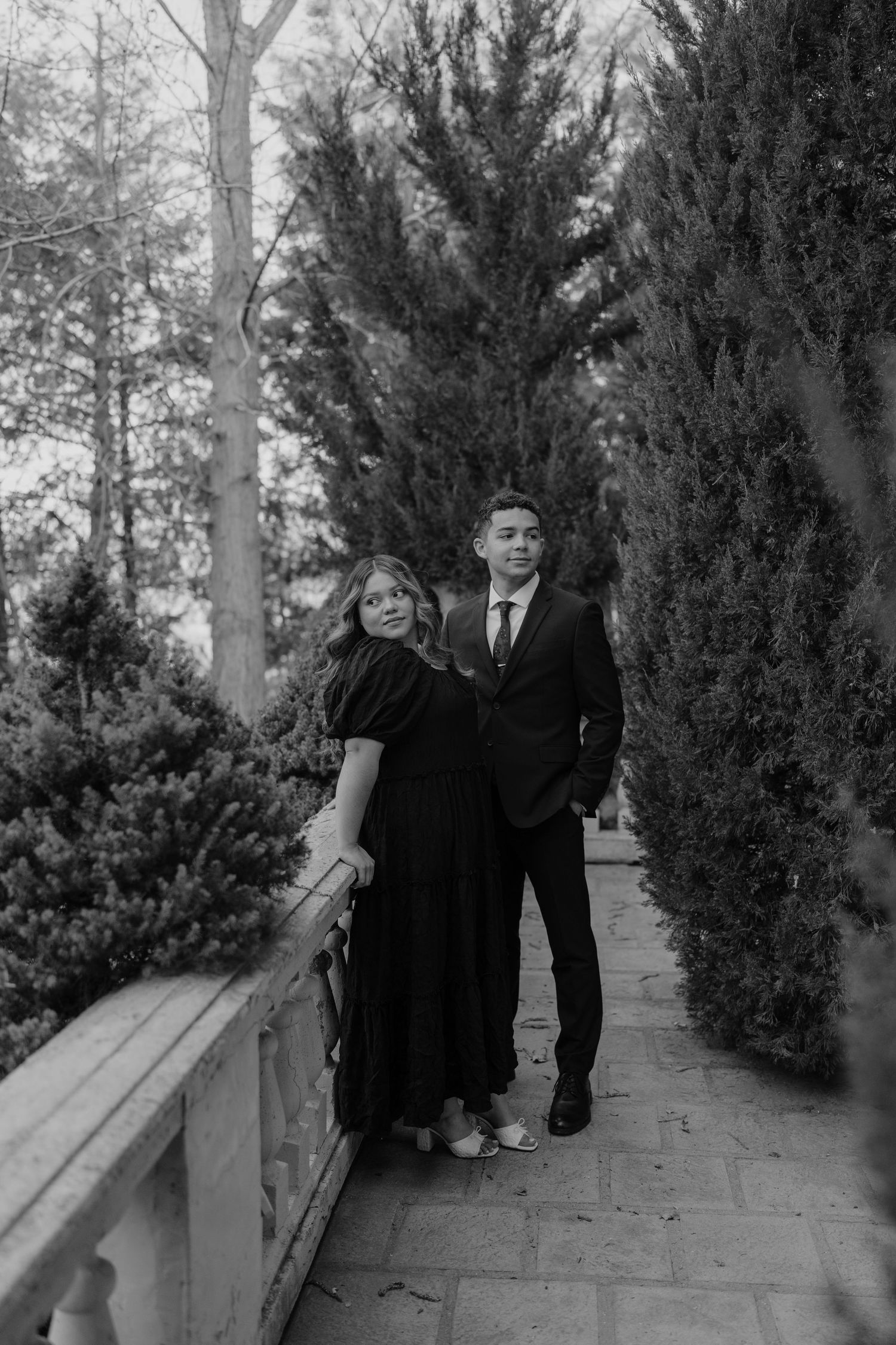 Black and white portrait of elegant couple in black attire posing along a garden stone pathway at La Caille Restaurant in Utah.