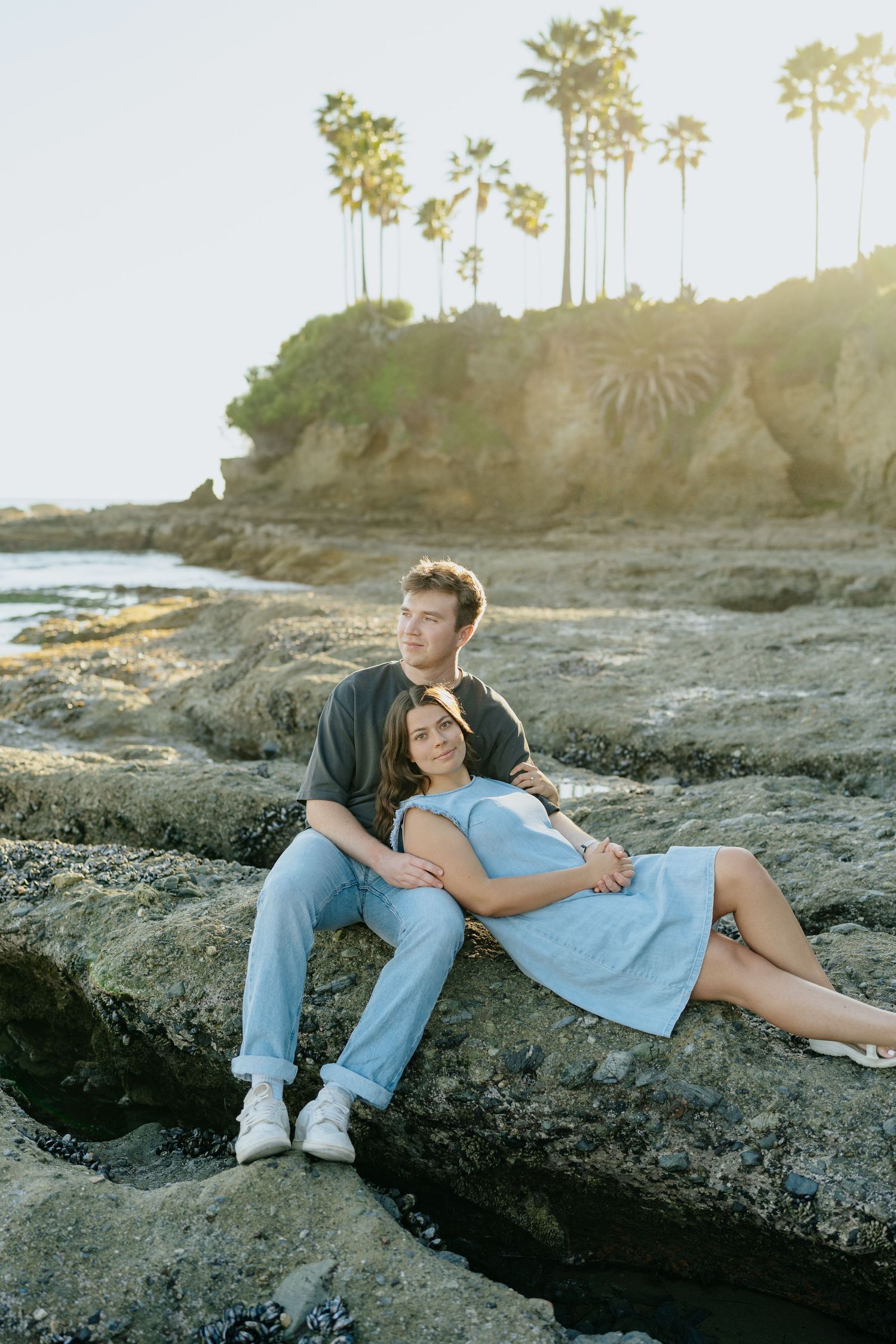 Couple sitting together on coastal rocks with palm trees and golden sunset behind them in Laguna Beach, California.