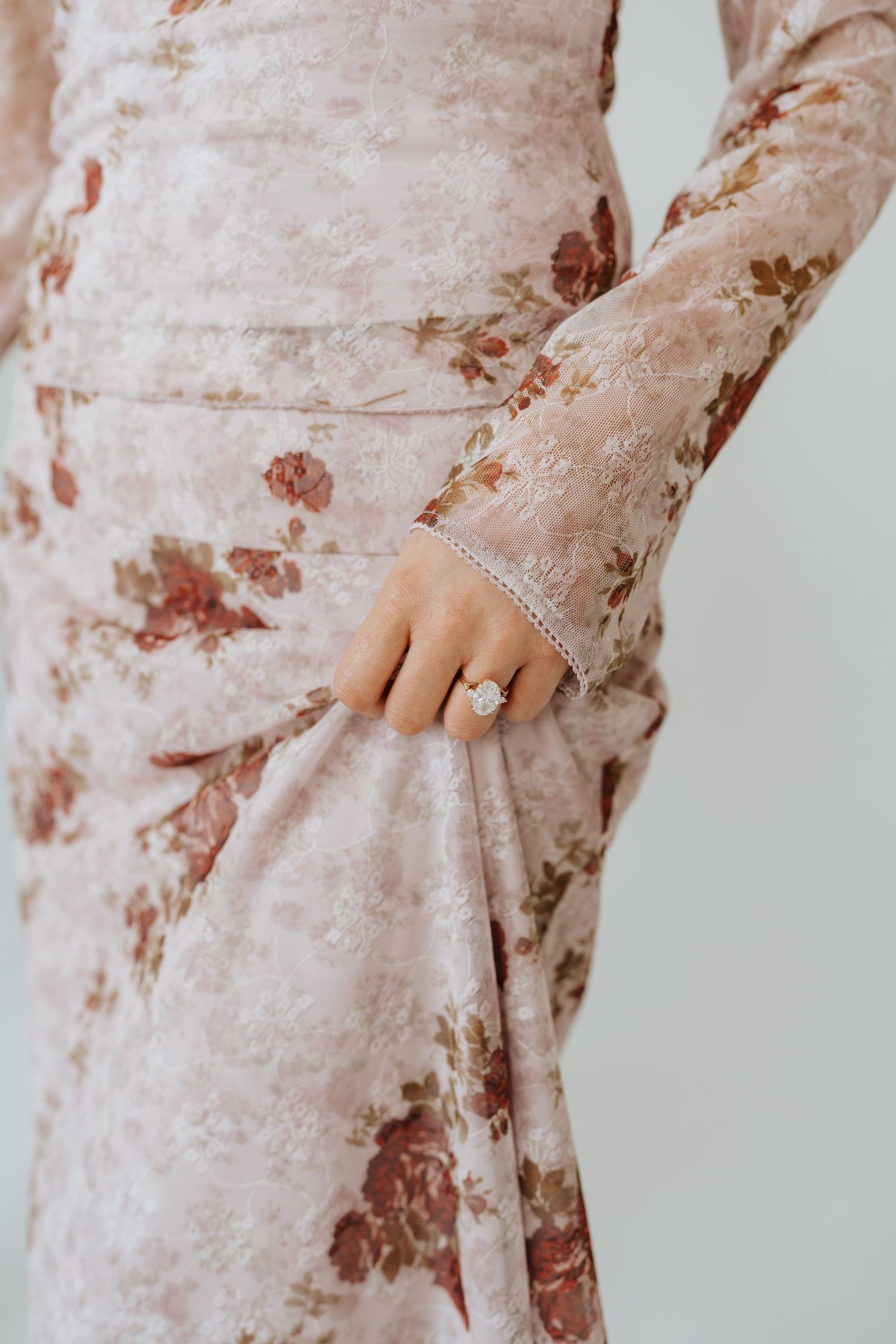 Close-up of womans hand wearing oval diamond engagement ring on floral lace dress.