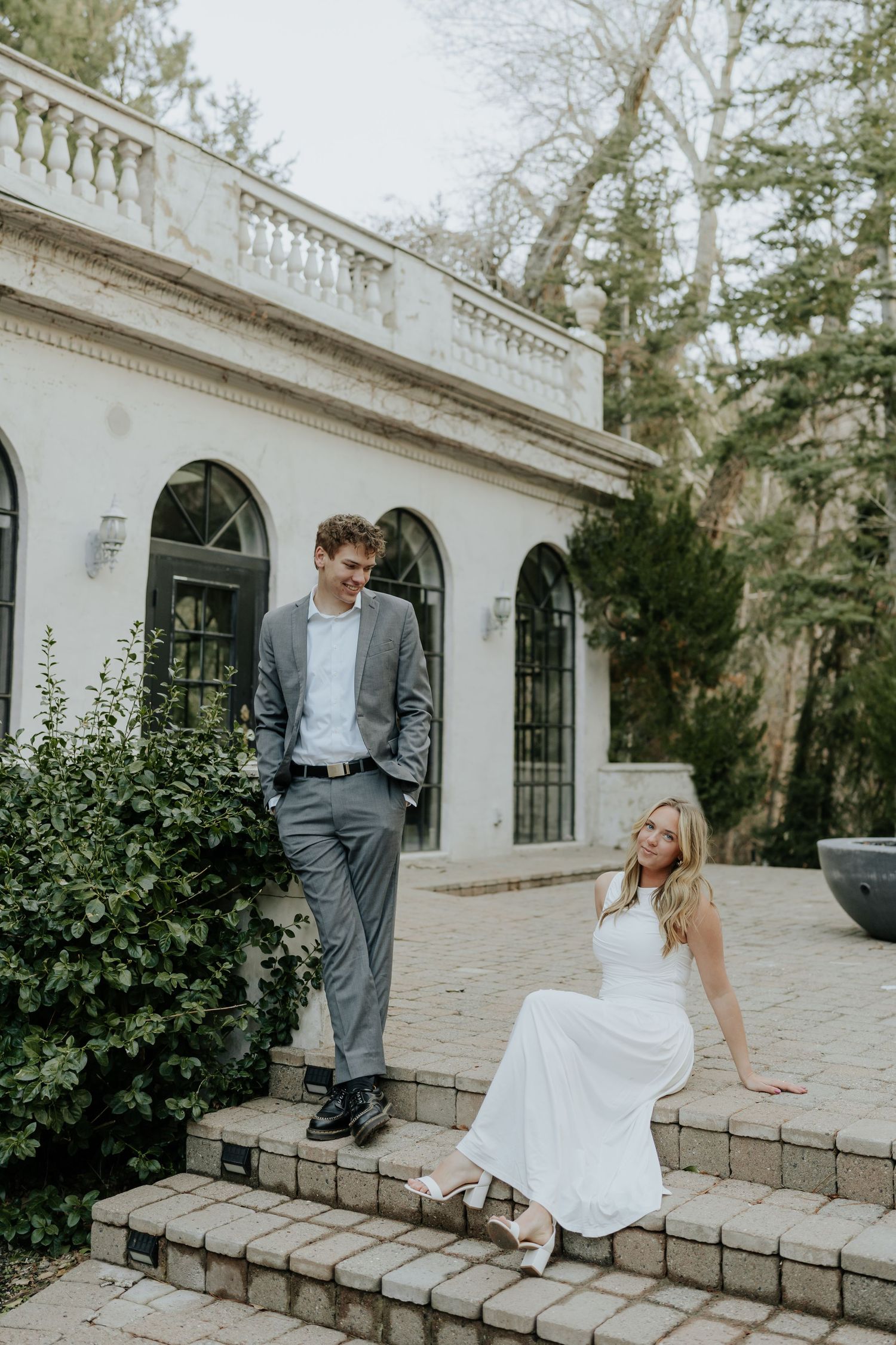 Couple posing on stone steps outside a white European-style villa, man standing, woman seated smiling at La Caille Restaurant in Utah.