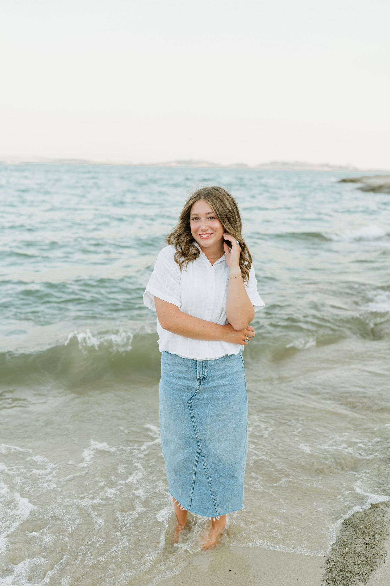 Smiling young woman in white top and denim skirt stands barefoot in ocean waves at sunset beach.