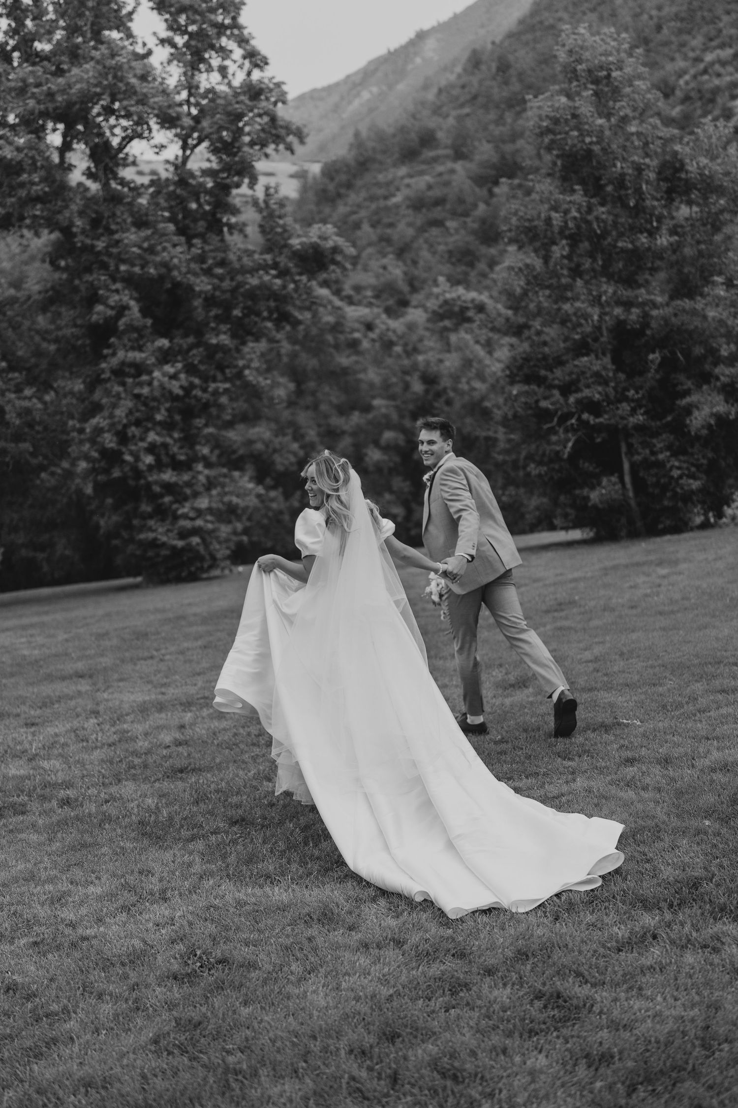 Black and white photo of playful bride and groom running on grass with mountain backdrop.