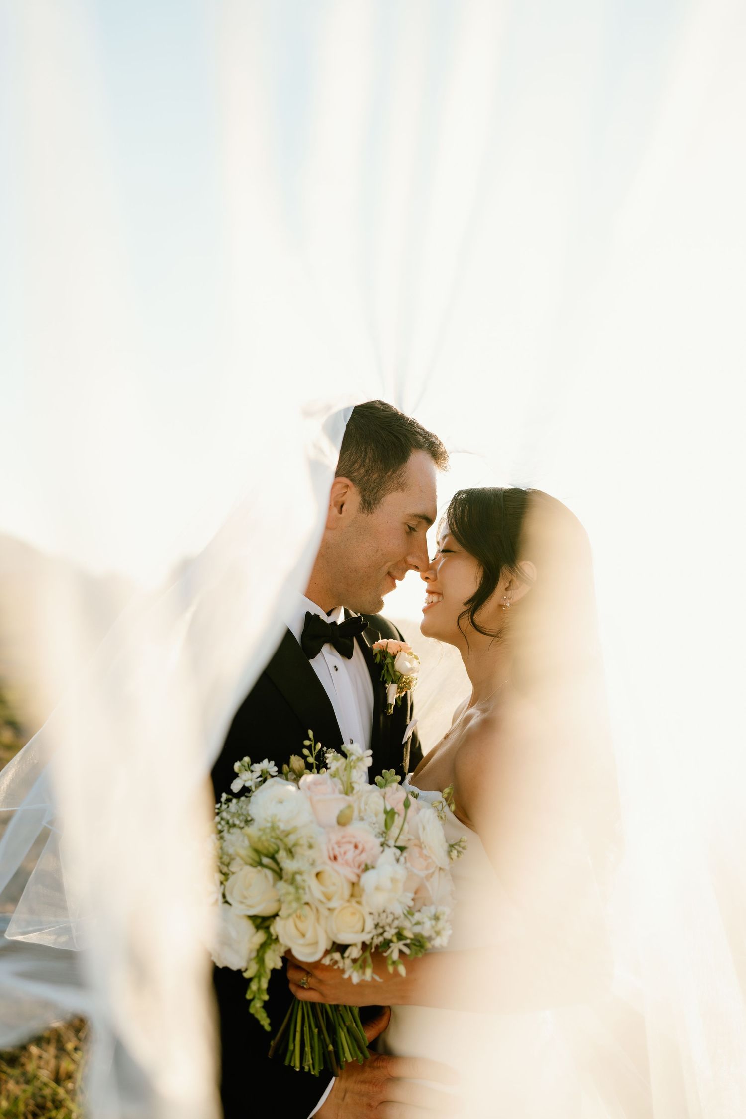 Bride and groom share a romantic forehead touch framed by a flowing veil, bride holding white floral bouquet.